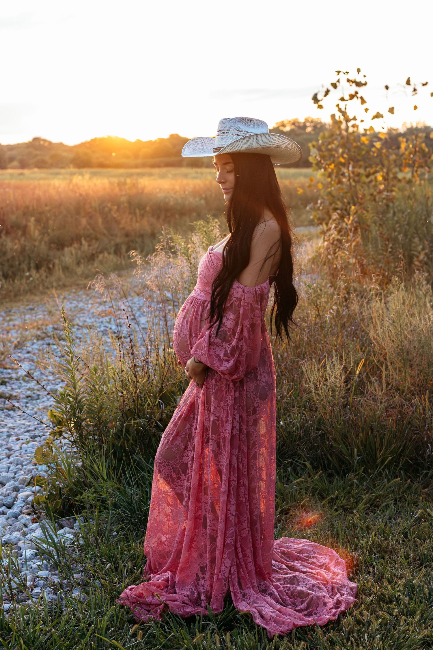 Maternity photo of woman in flowing pink lace dress and cowboy hat at golden hour outdoors.