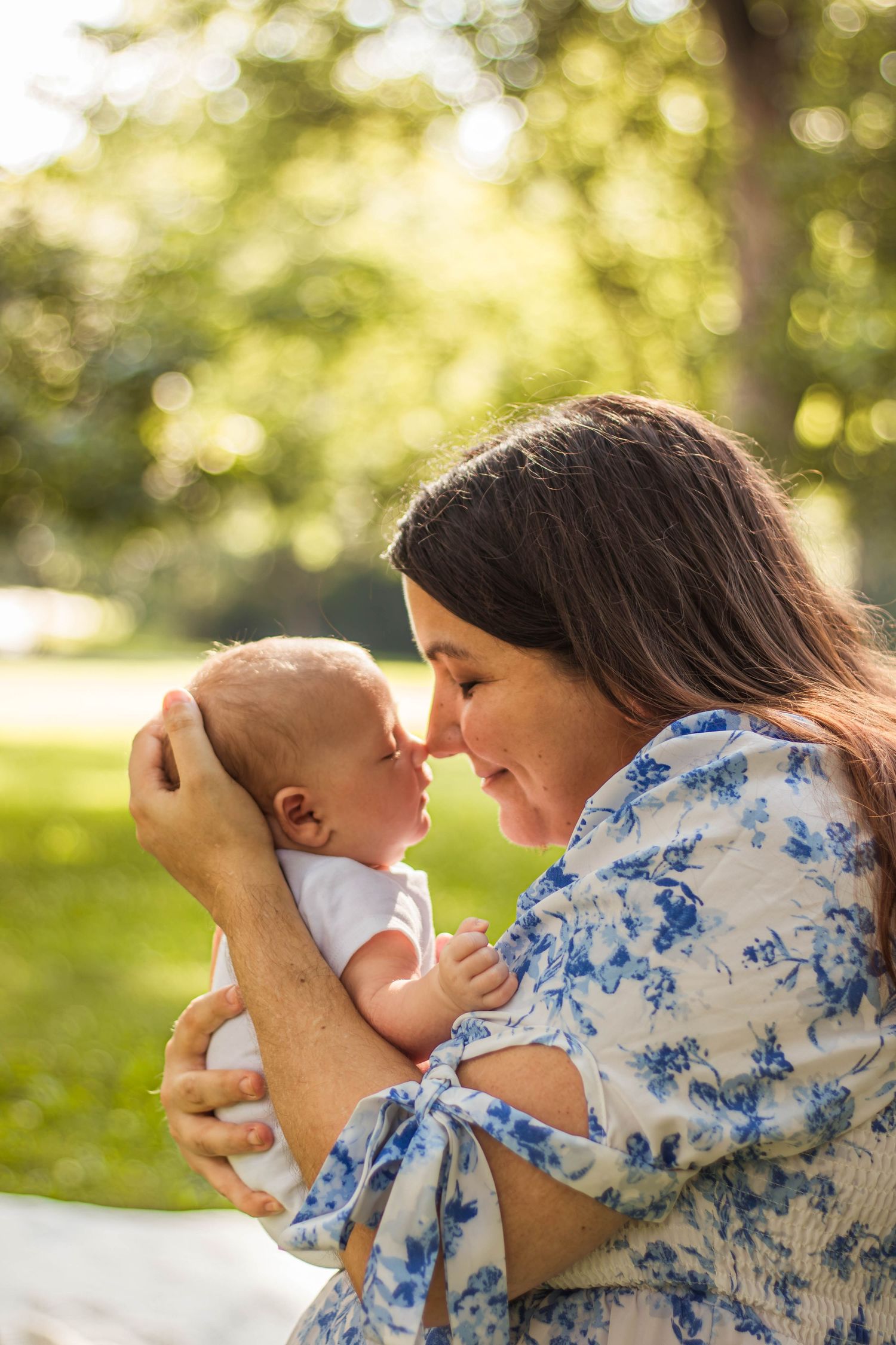 Loving mother touching noses with newborn baby while sitting outdoors in sunny green park.