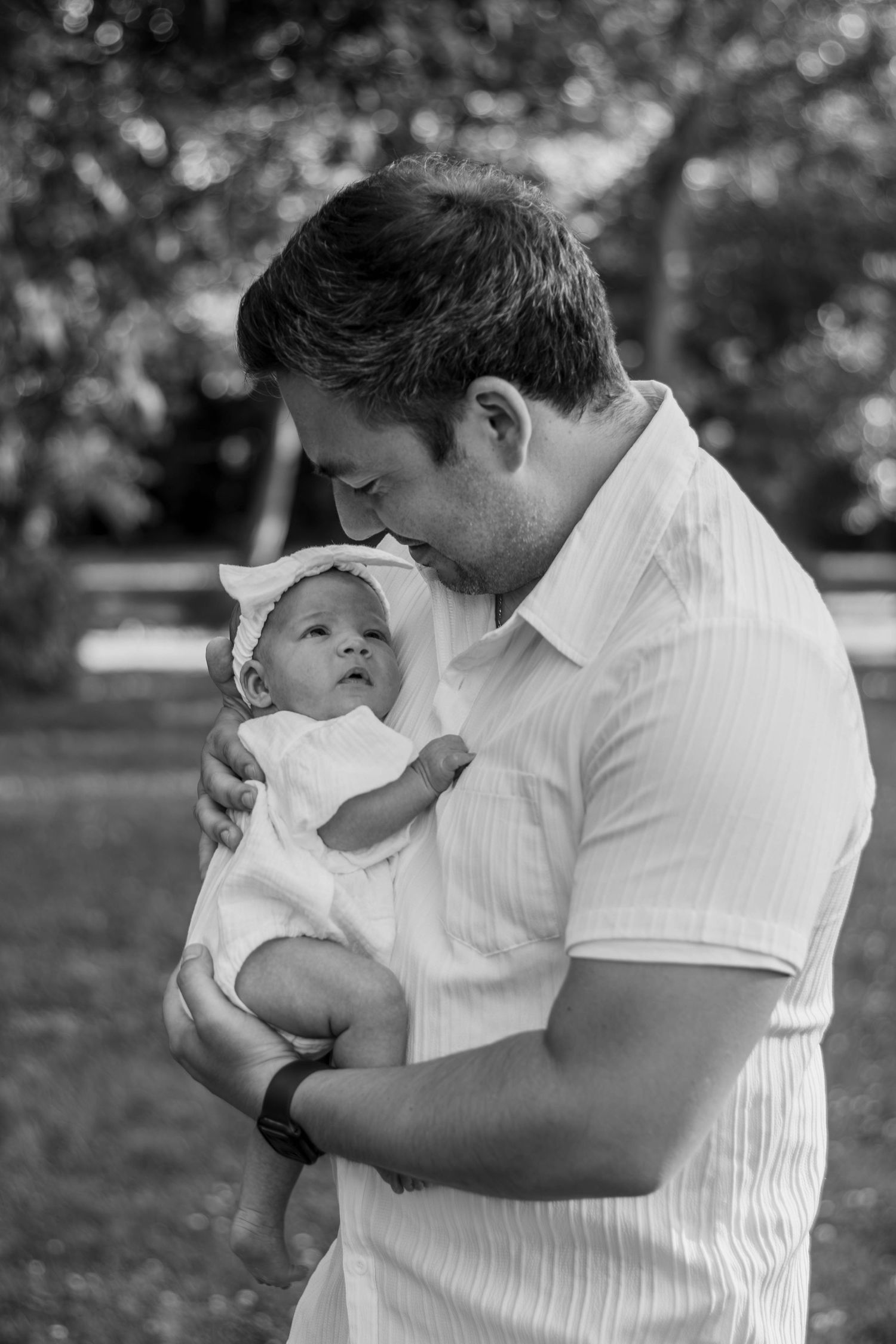 Black and white photo of father holding newborn baby outdoors in park.