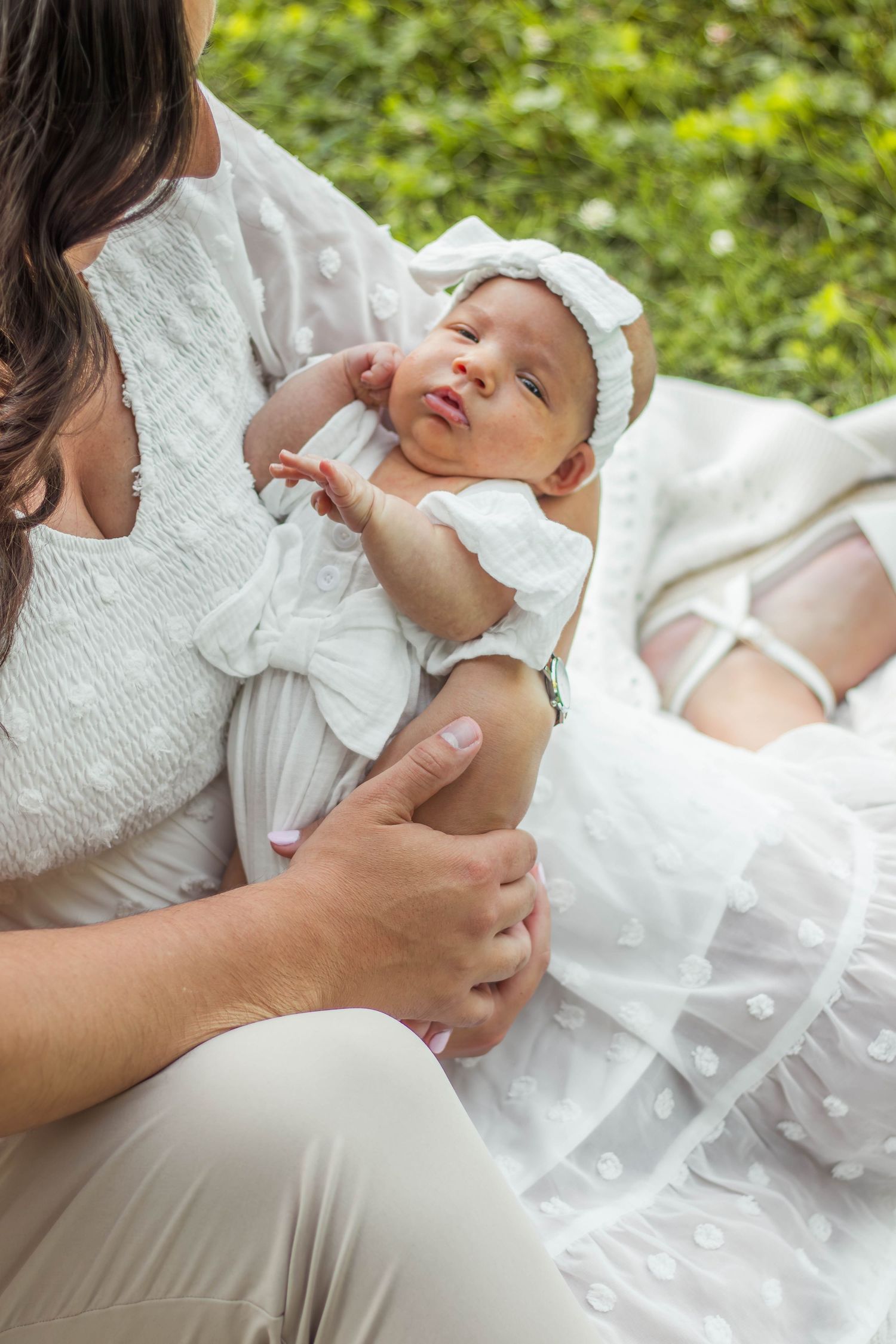 Mother in white dress holding newborn baby with white bow headband outdoors.