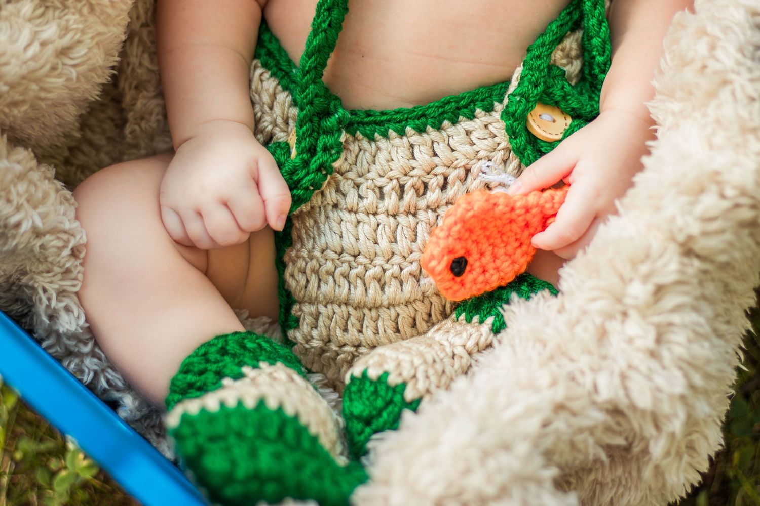 Newborn baby in crochet fishing outfit holding orange fish toy while resting on fluffy blanket.