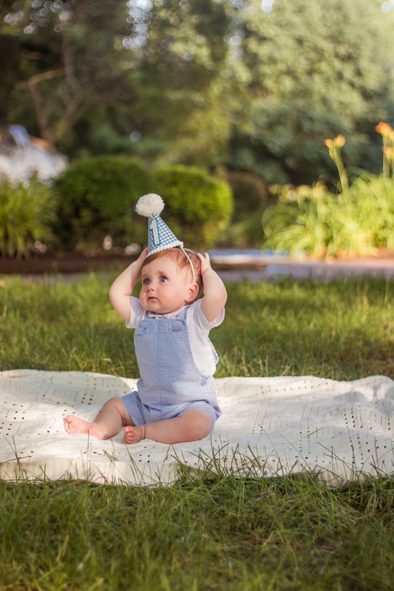 Adorable baby in blue romper touching party hat while sitting on blanket in garden.
