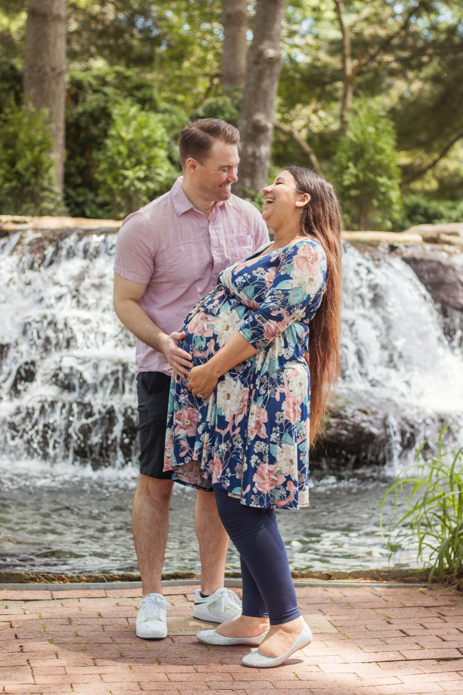 Happy couple embracing in front of a waterfall, woman wearing floral dress showing baby bump.