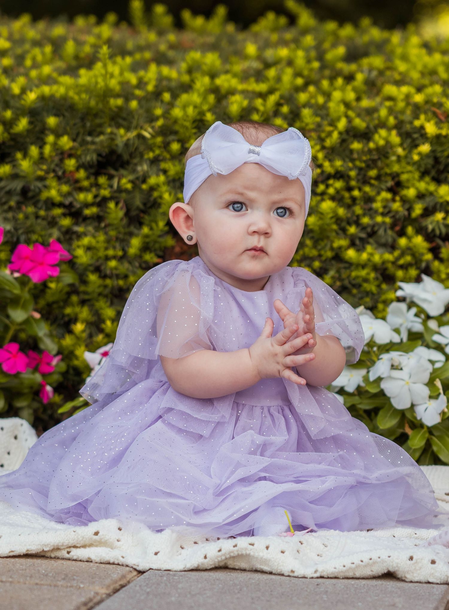Elegant baby girl in lavender tulle dress with white bow headband clapping near garden flowers.