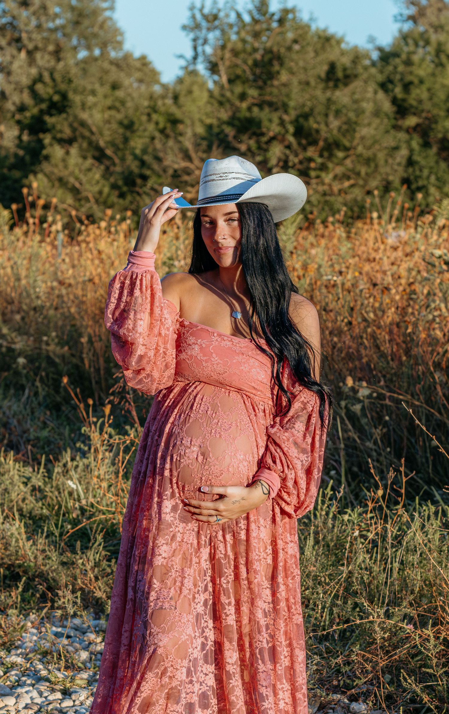 Pregnant woman in pink lace dress and cowboy hat holding belly in autumn field at sunset.