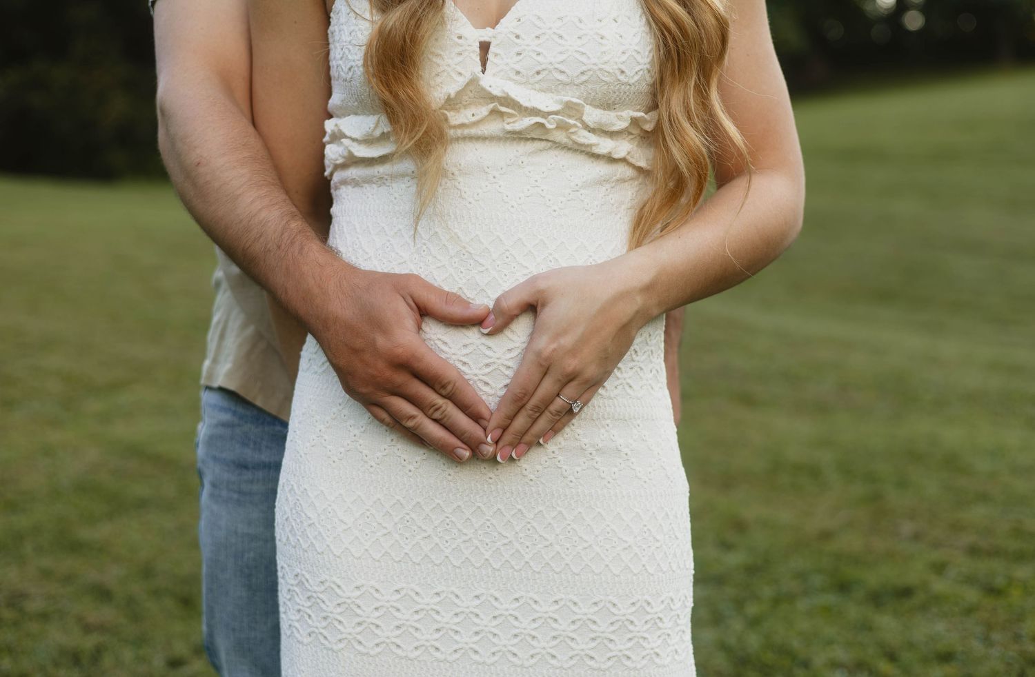 Close-up of couple forming heart shape with hands on pregnant belly in white lace maternity dress.