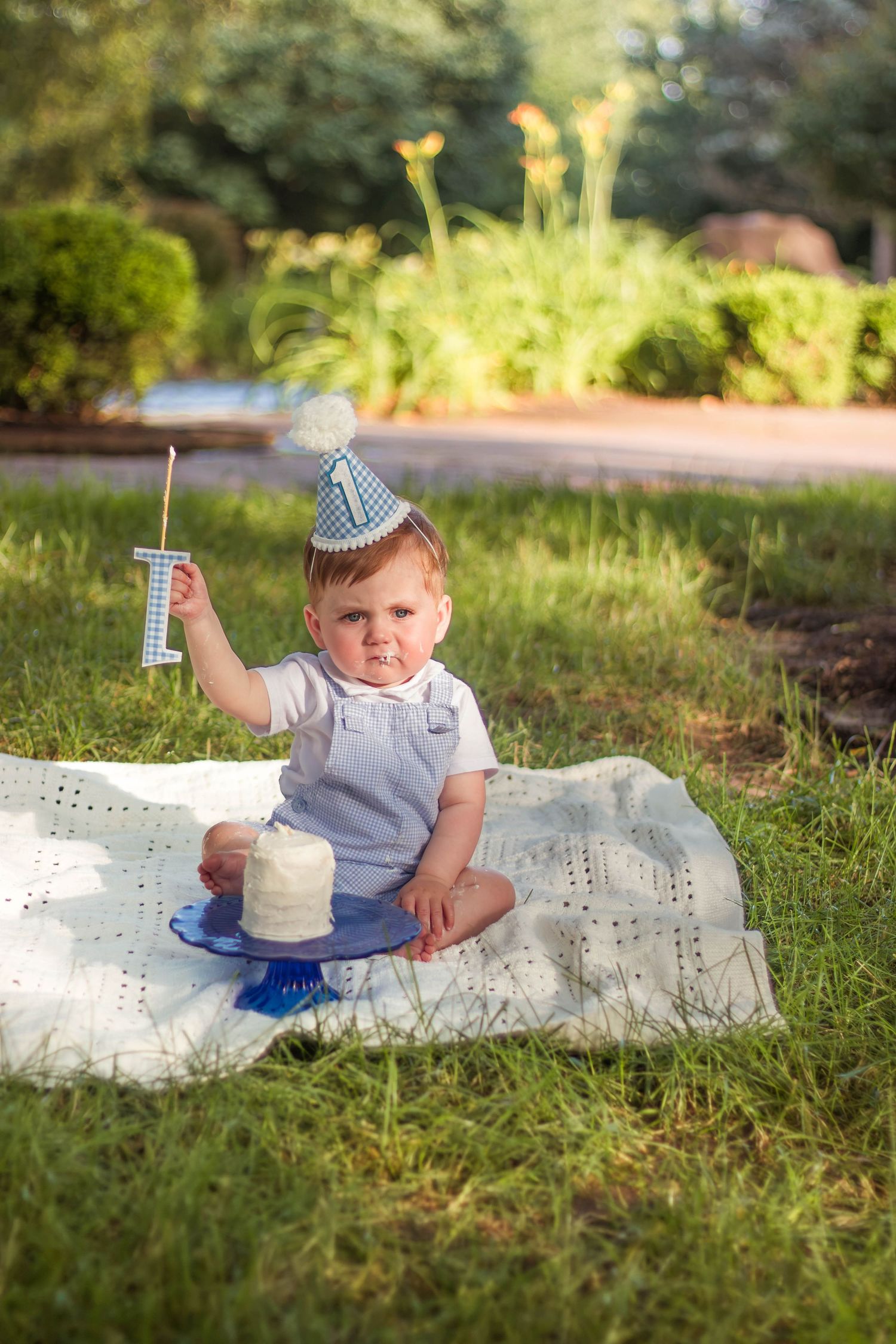 Baby in blue outfit holding number one candle beside smash cake during first birthday celebration.
