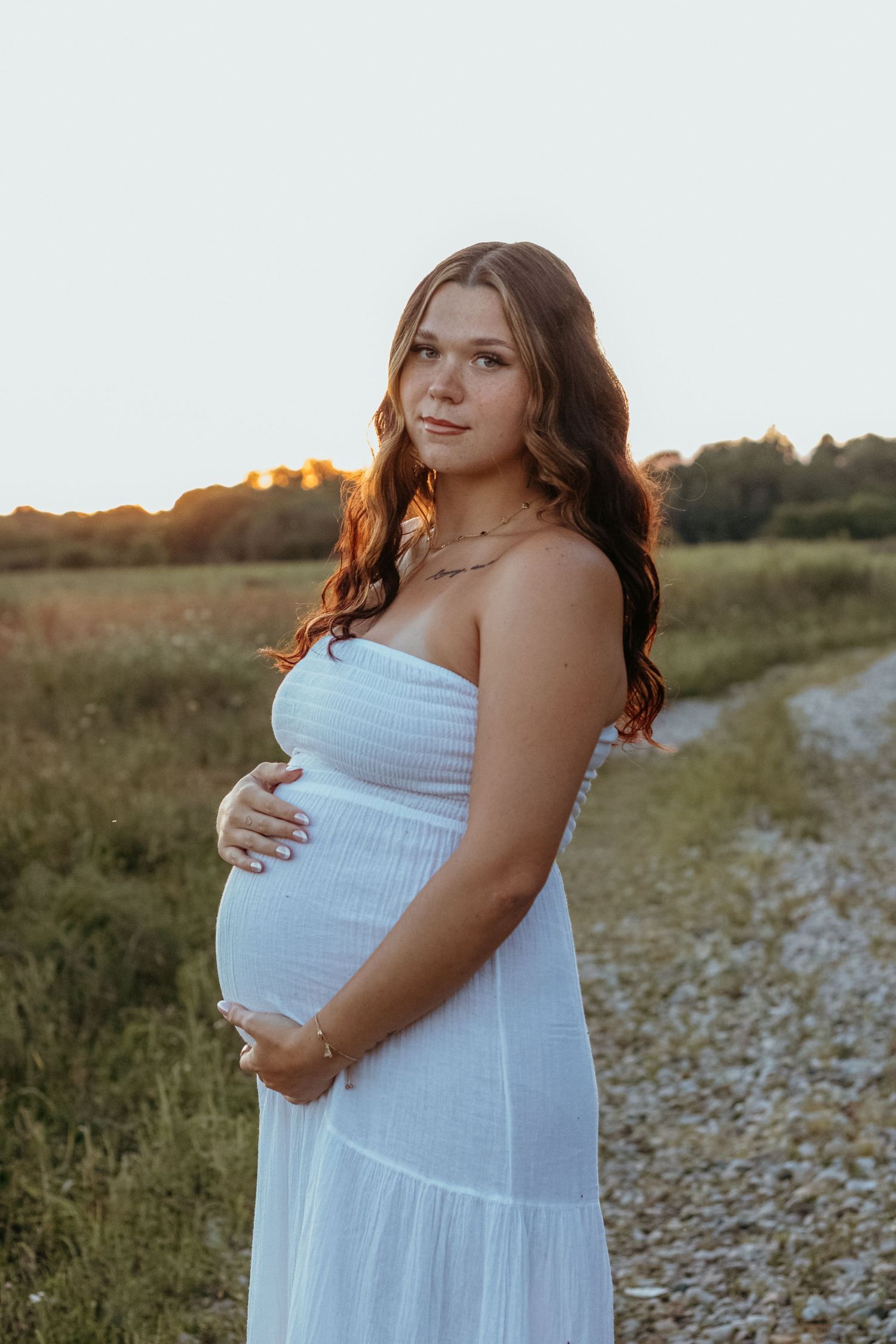 Expectant mother in white dress holding baby bump during sunset maternity photoshoot outdoors.