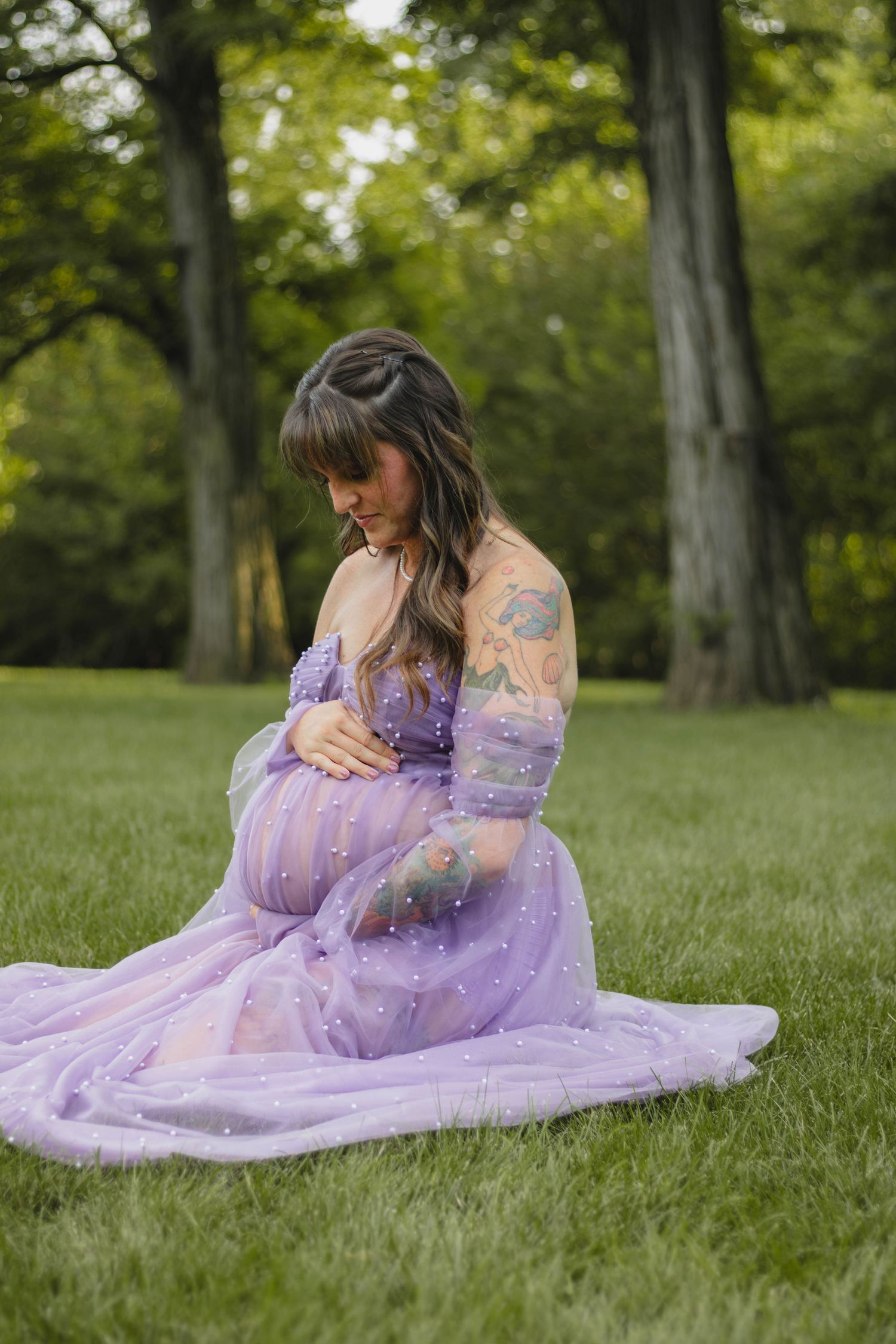 Pregnant woman in lavender tulle maternity gown sitting on grass in a lush green park.