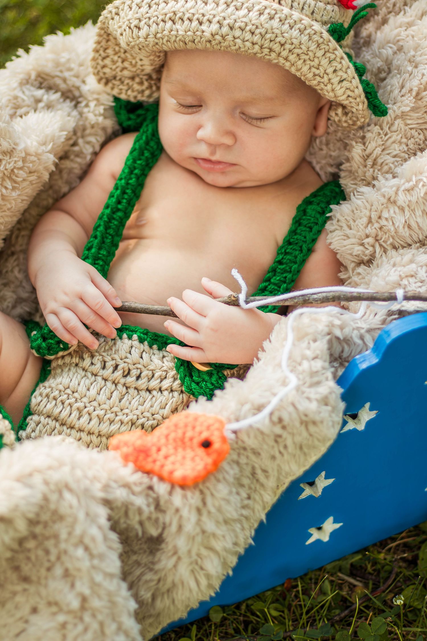 Sleeping newborn dressed in crochet fisherman outfit holding tiny fishing rod with orange fish.