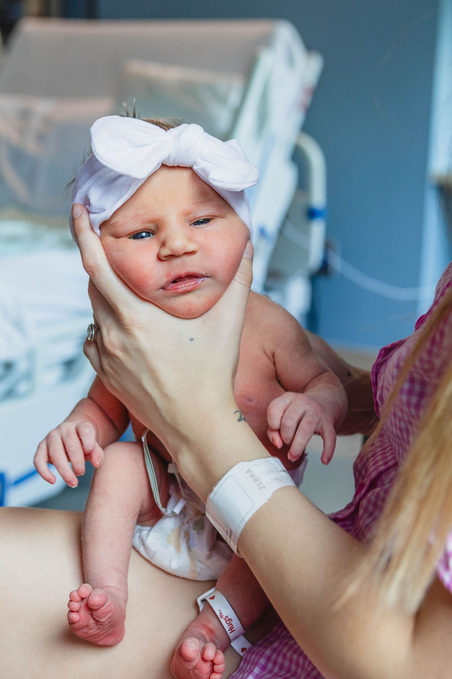 Newborn baby girl with white bow headband being held by mother in hospital room.