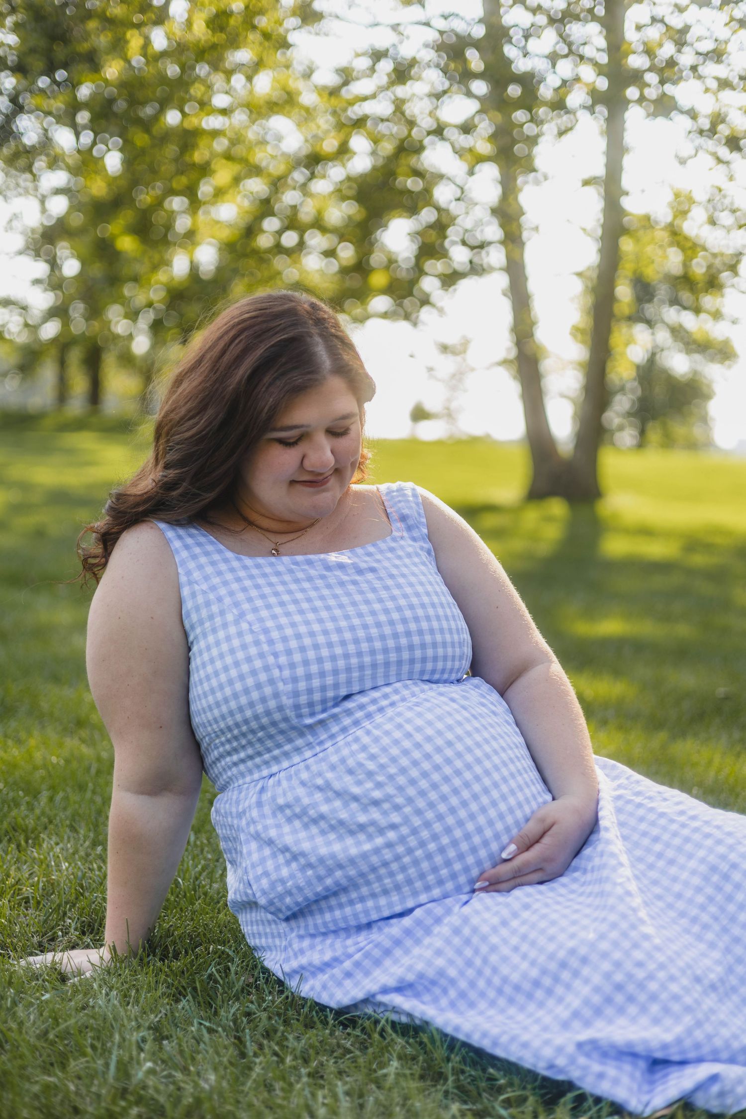 Pregnant woman in blue gingham dress sitting on grass looking down at belly in sunny park.