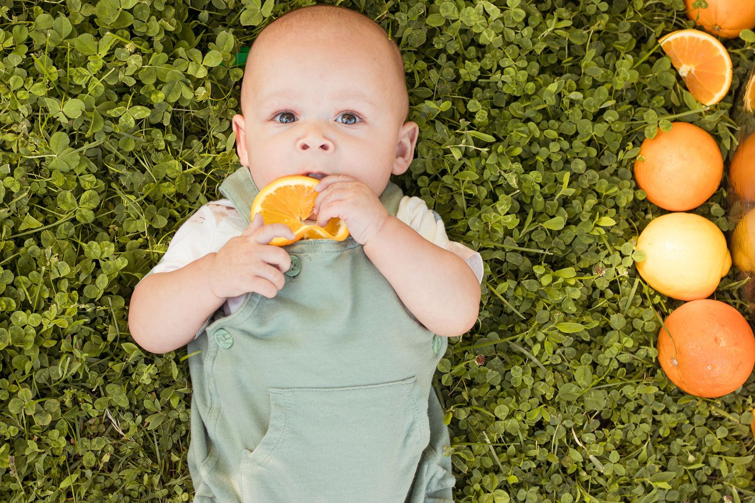 Cute baby in green romper lying on grass holding an orange slice to mouth.
