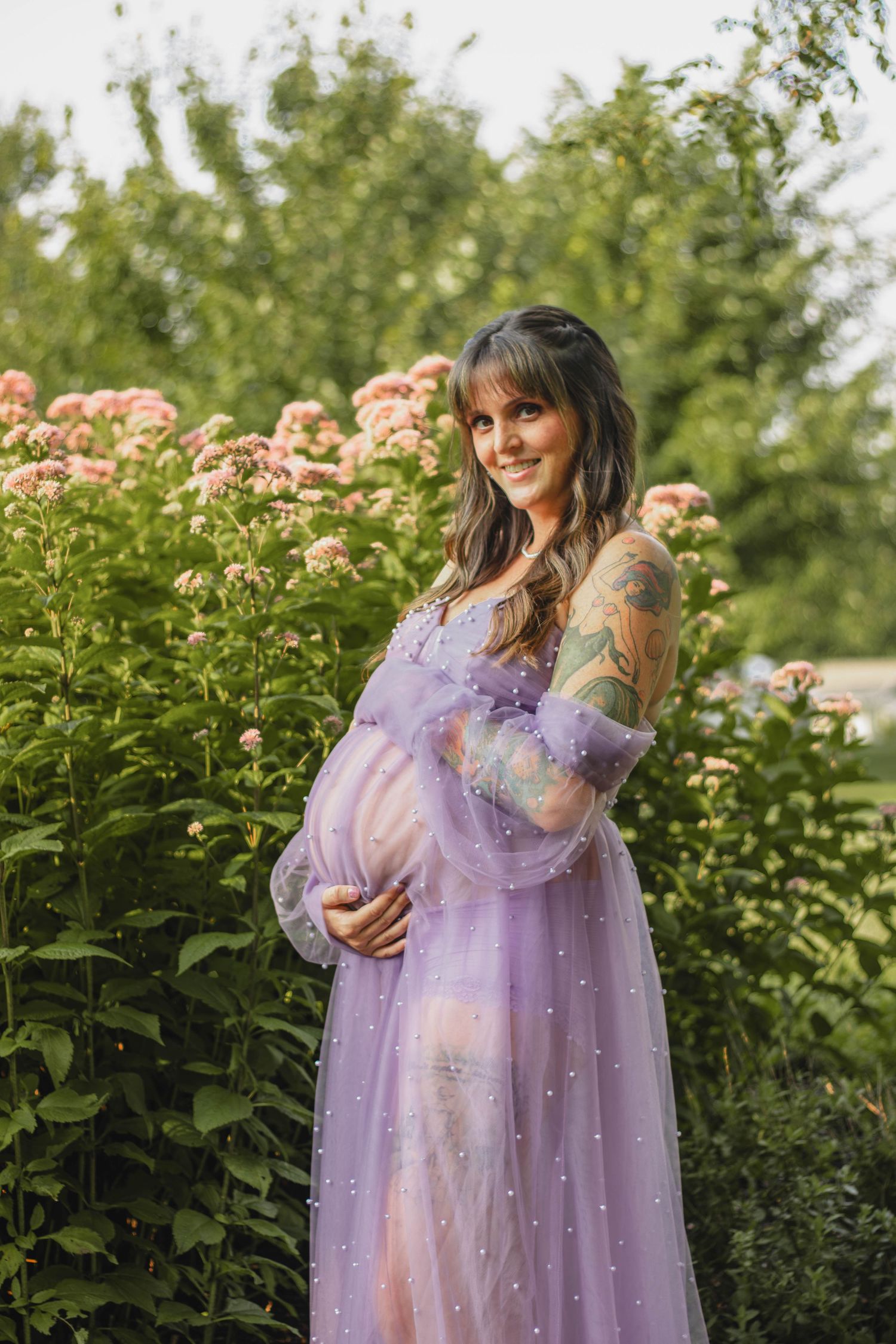 Pregnant woman in sheer lilac pearl-embellished gown standing by pink flowering bushes.