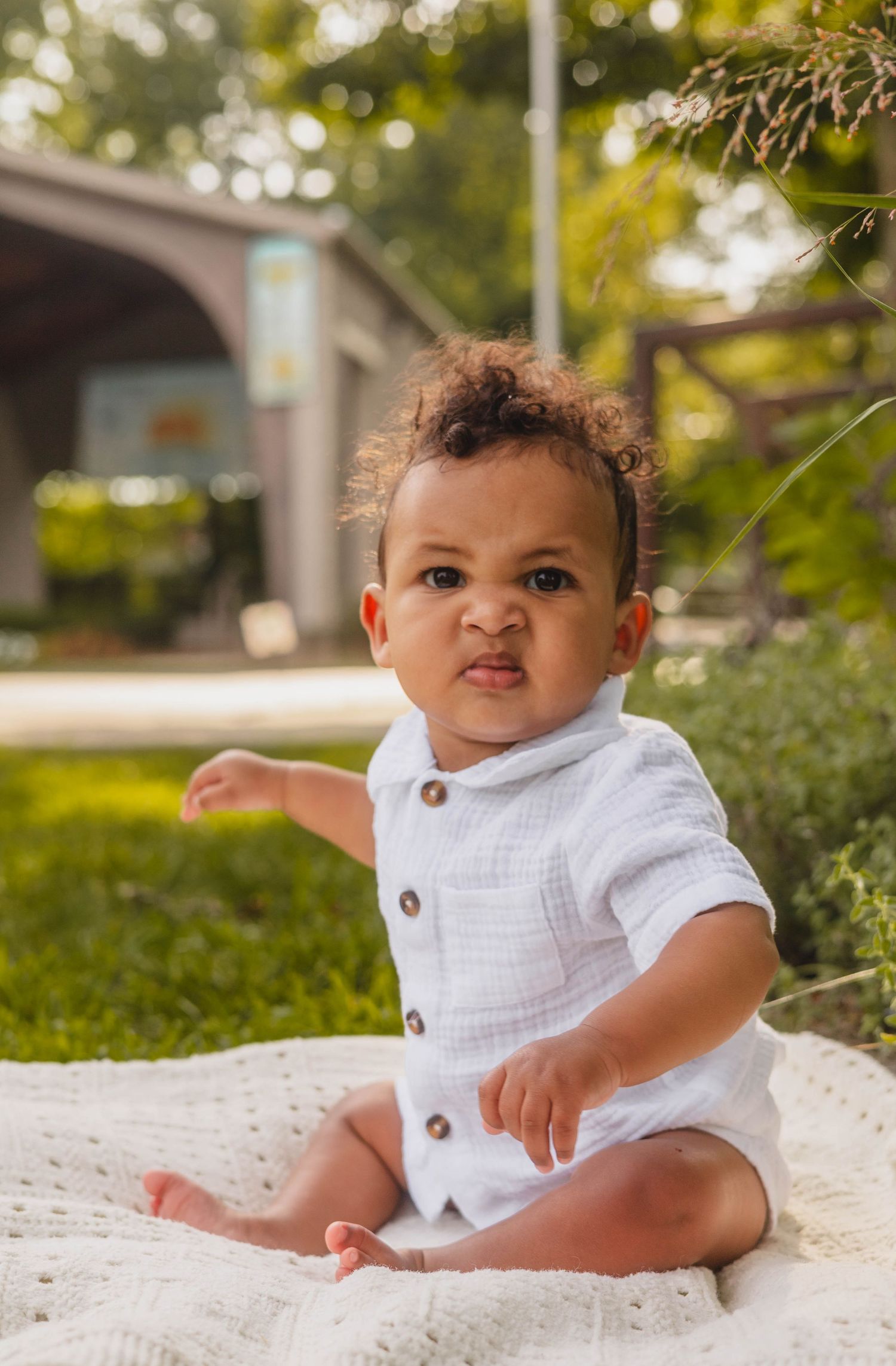 Adorable baby boy in white button romper sitting on blanket outdoors.