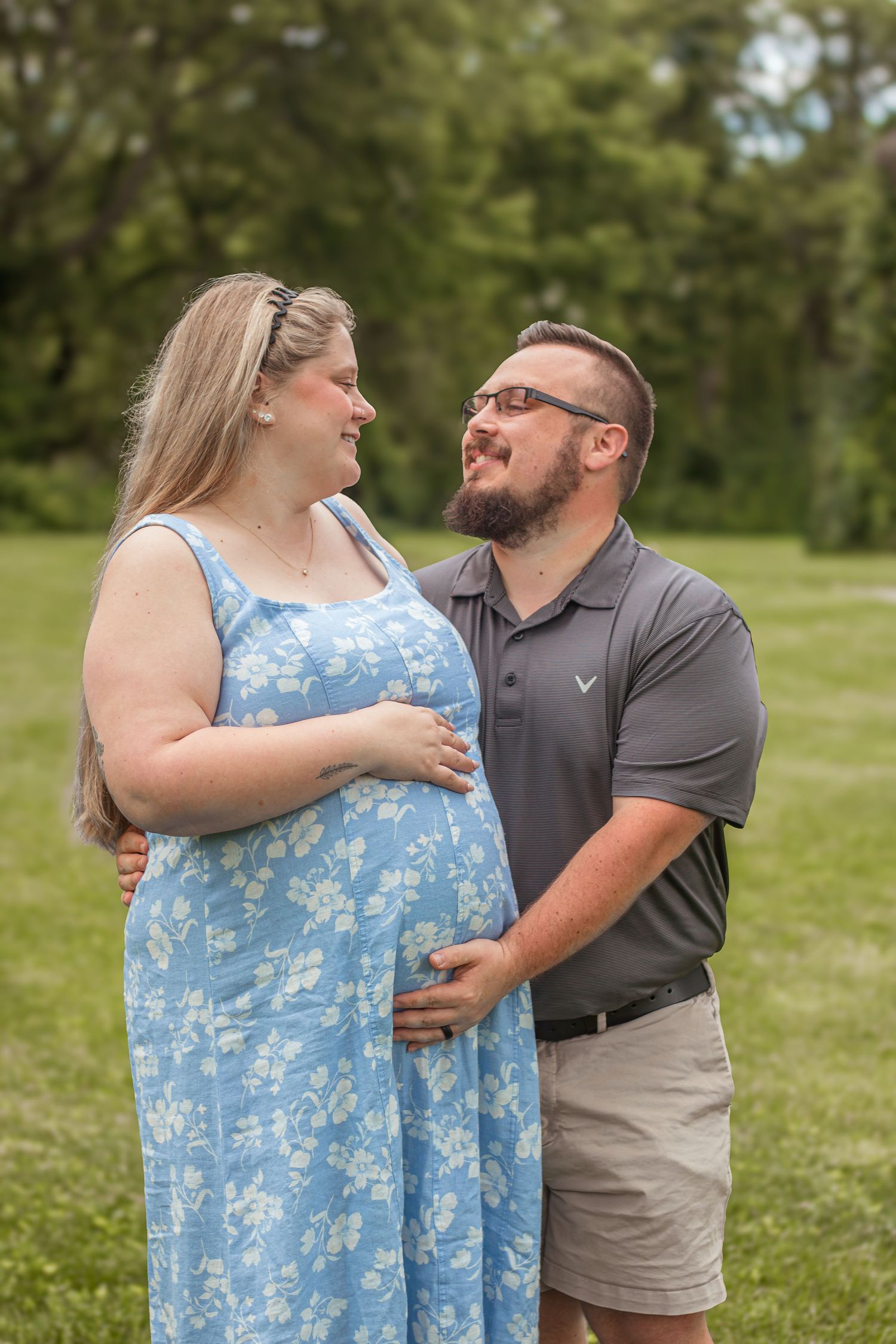 Pregnant woman in blue floral dress and bearded partner smiling at each other in park.