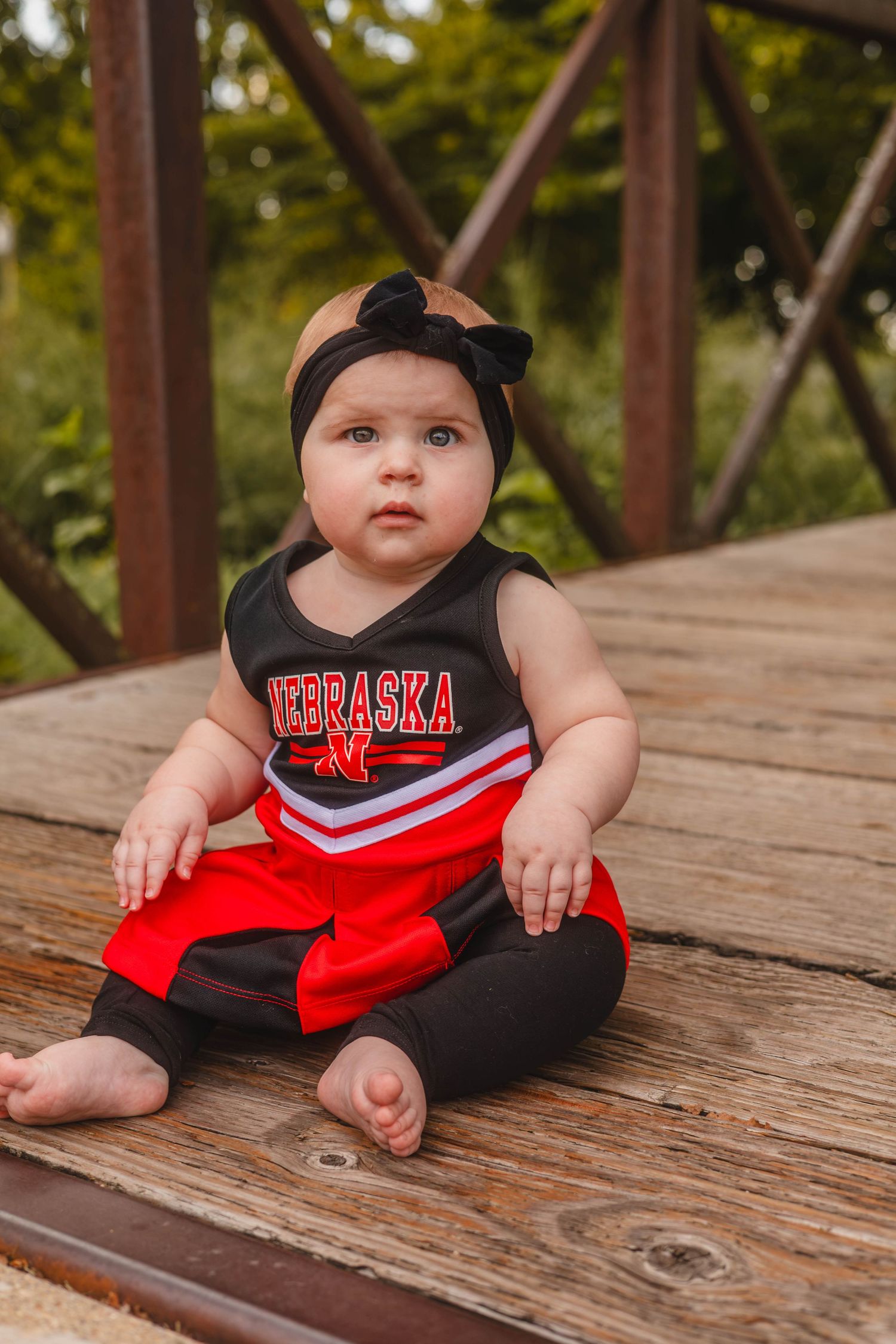 Adorable baby in Nebraska Cornhuskers red and black cheerleader dress sitting on wooden deck outside.