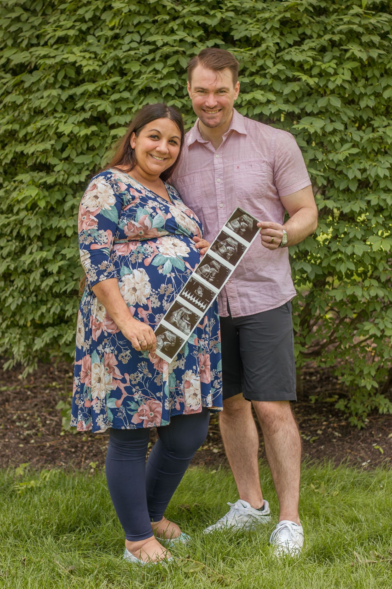 Smiling pregnant couple holding ultrasound photos outdoors in front of green ivy wall.