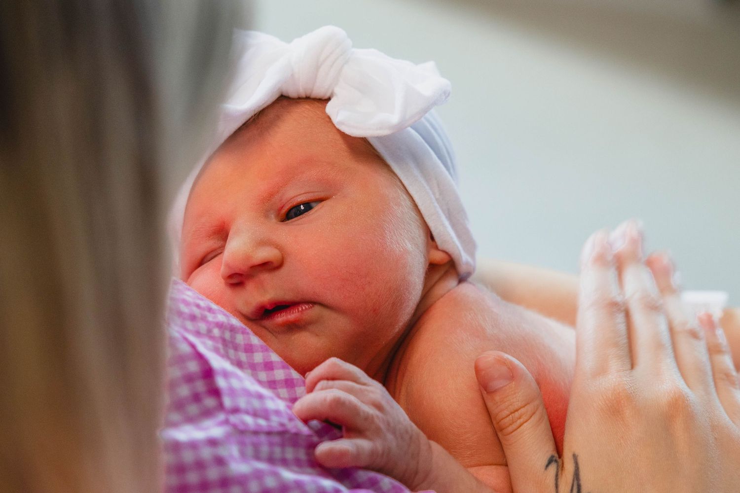Close-up of newborn baby with white bow headband resting in mothers arms.