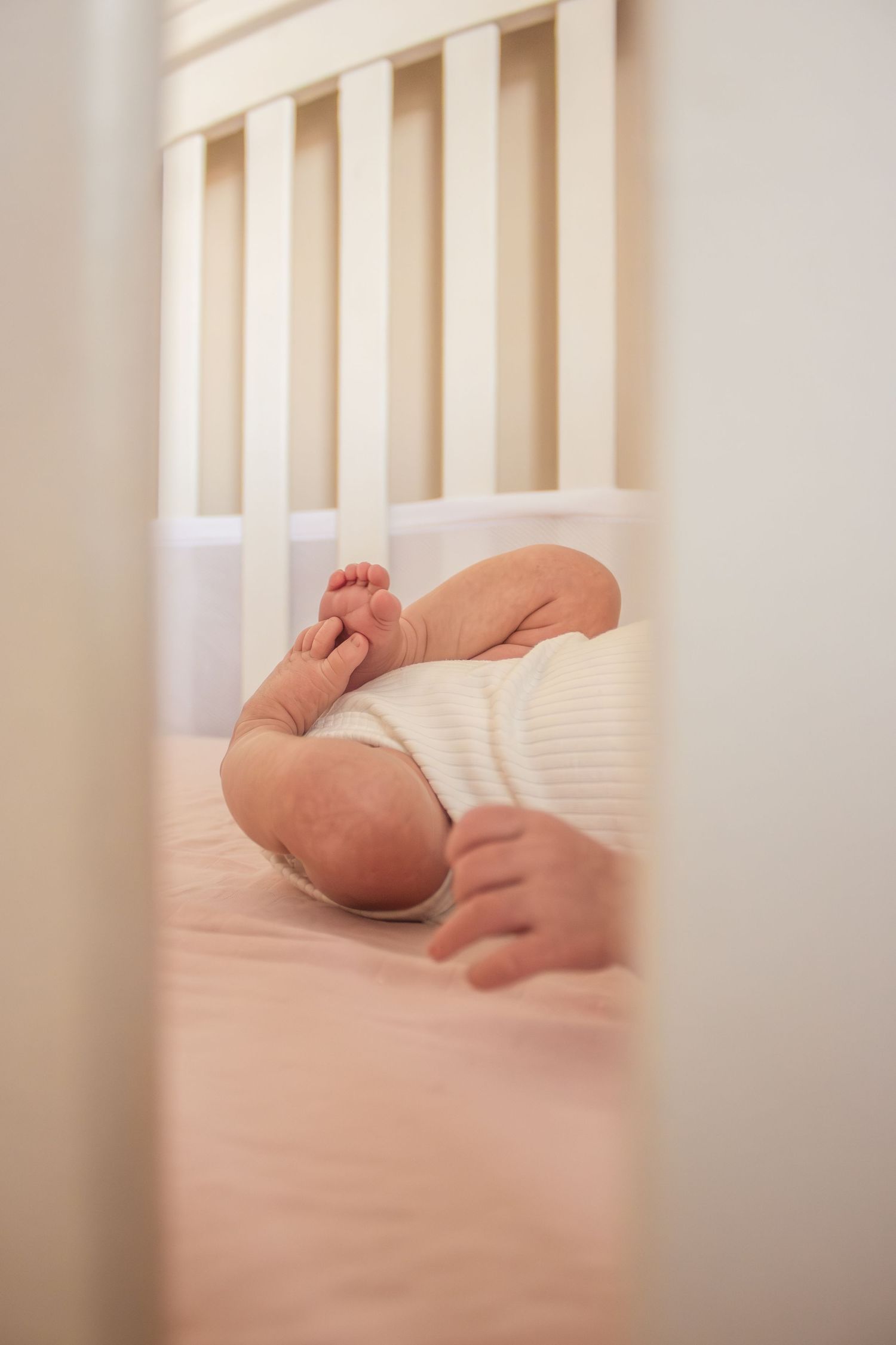 Peaceful newborn baby sleeping in white crib, wearing white onesie with tiny feet visible.