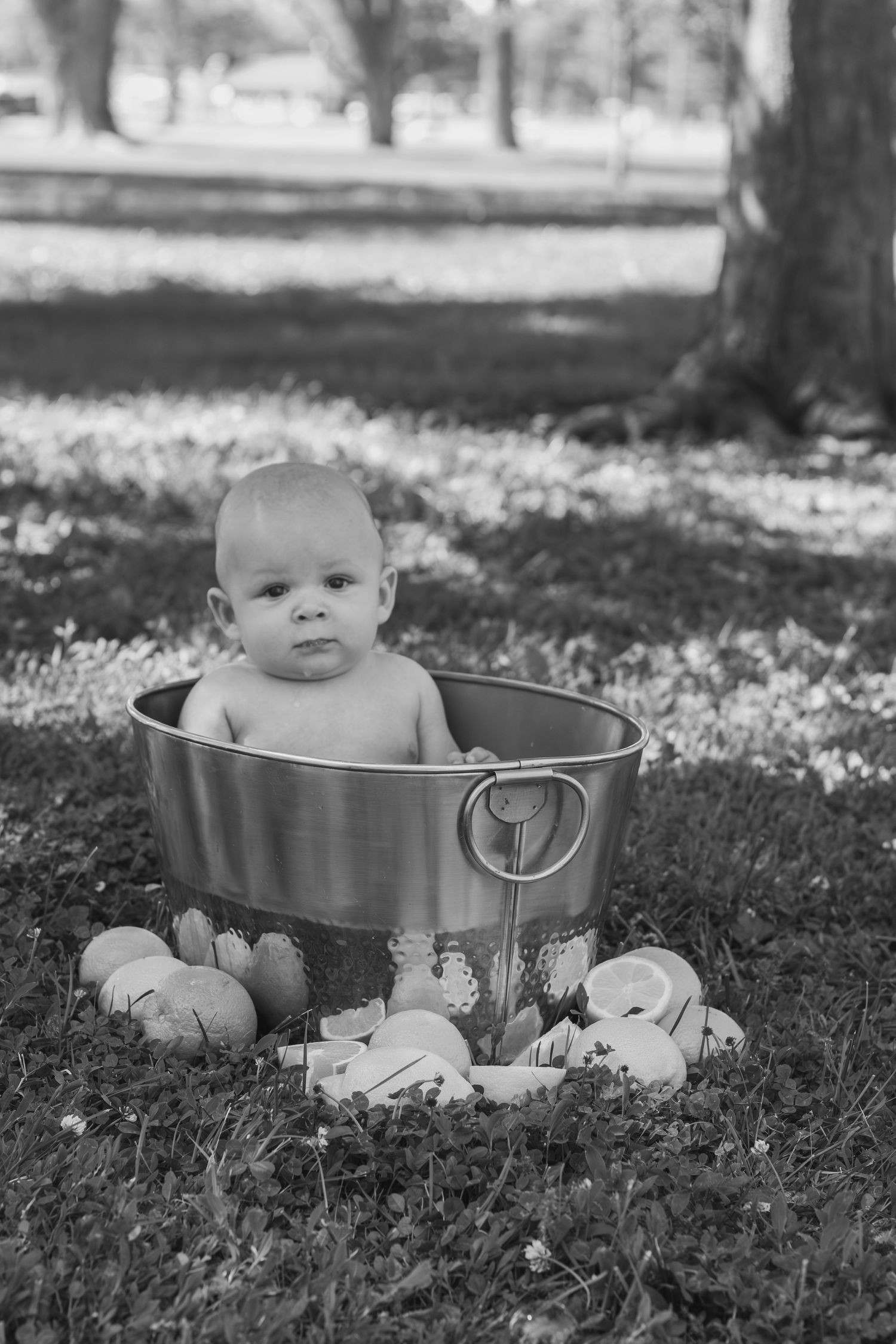 Black and white photo of a baby sitting in a metal tub outdoors surrounded by fruit.