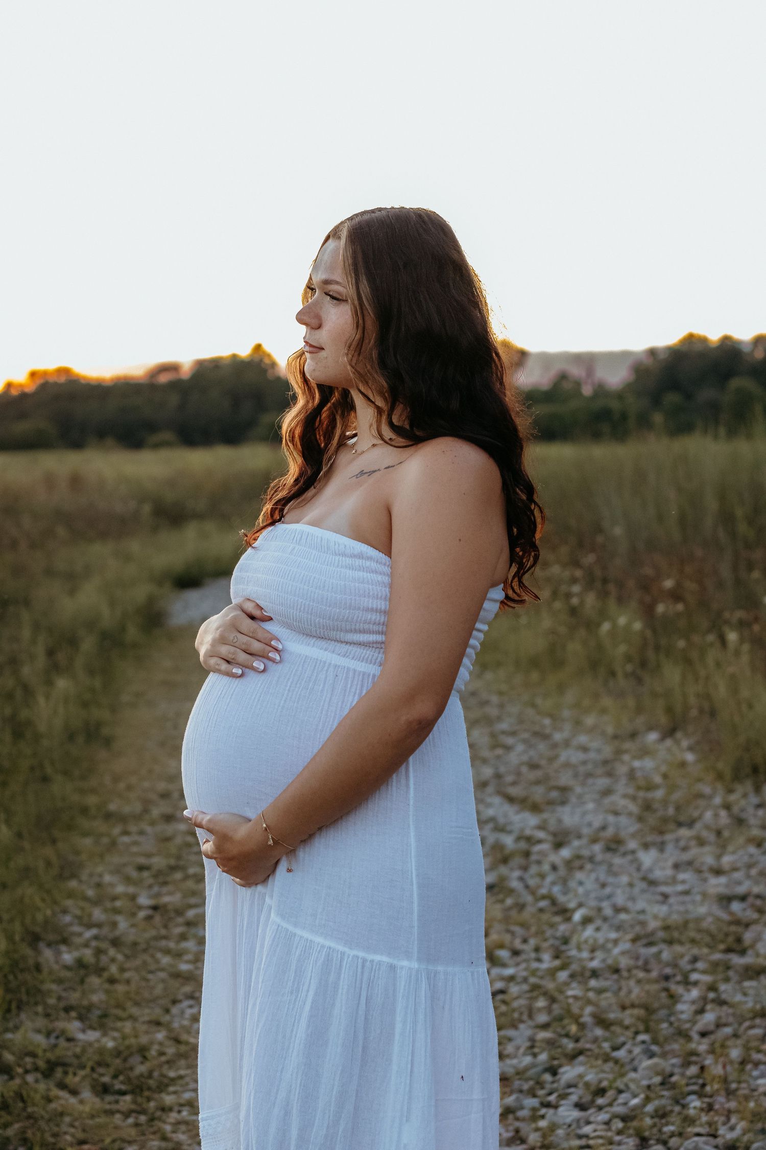 Pregnant woman in white strapless dress cradling baby bump at golden hour in a field.
