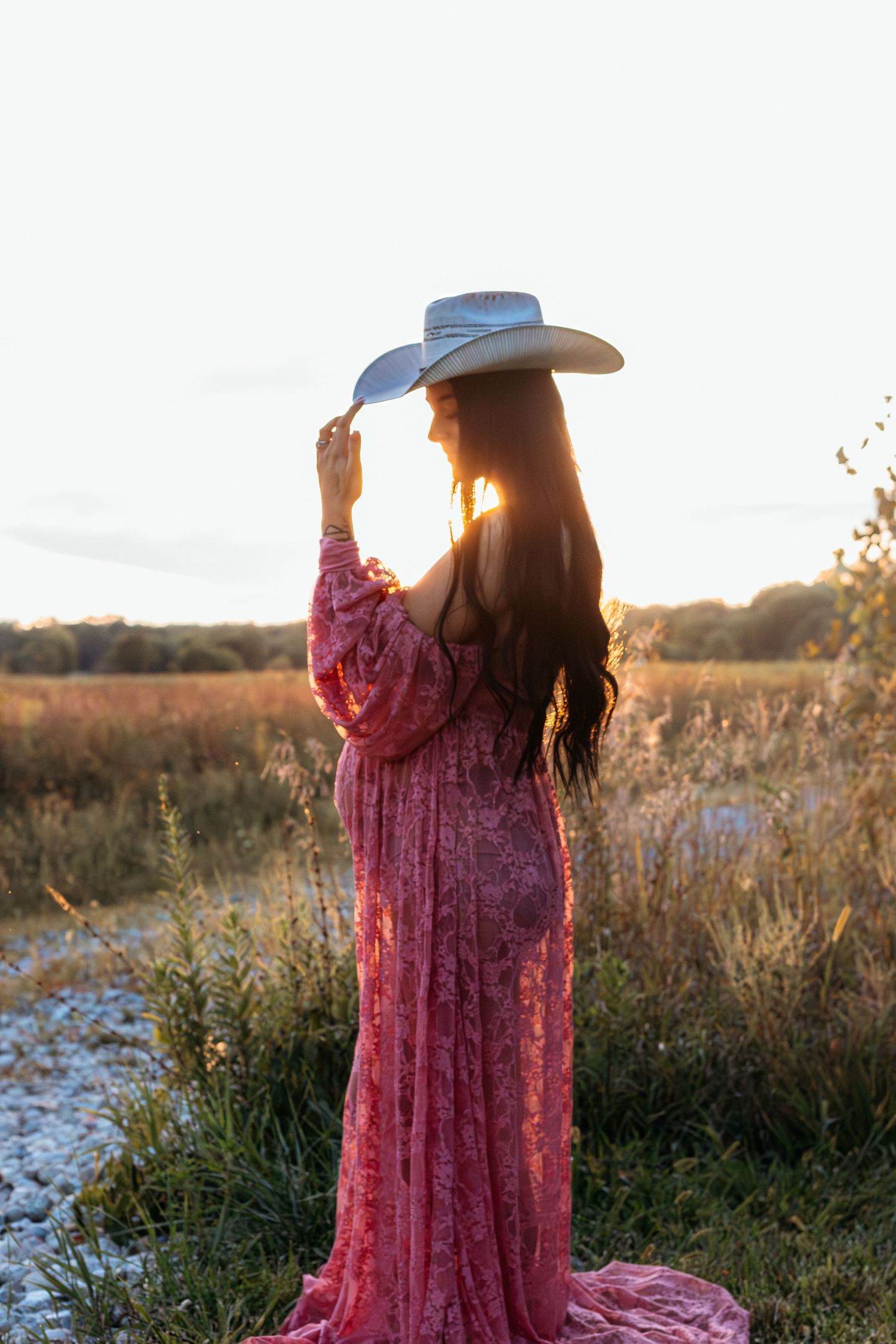 Pregnant woman in pink lace dress and white cowboy hat backlit by golden sunset in field.