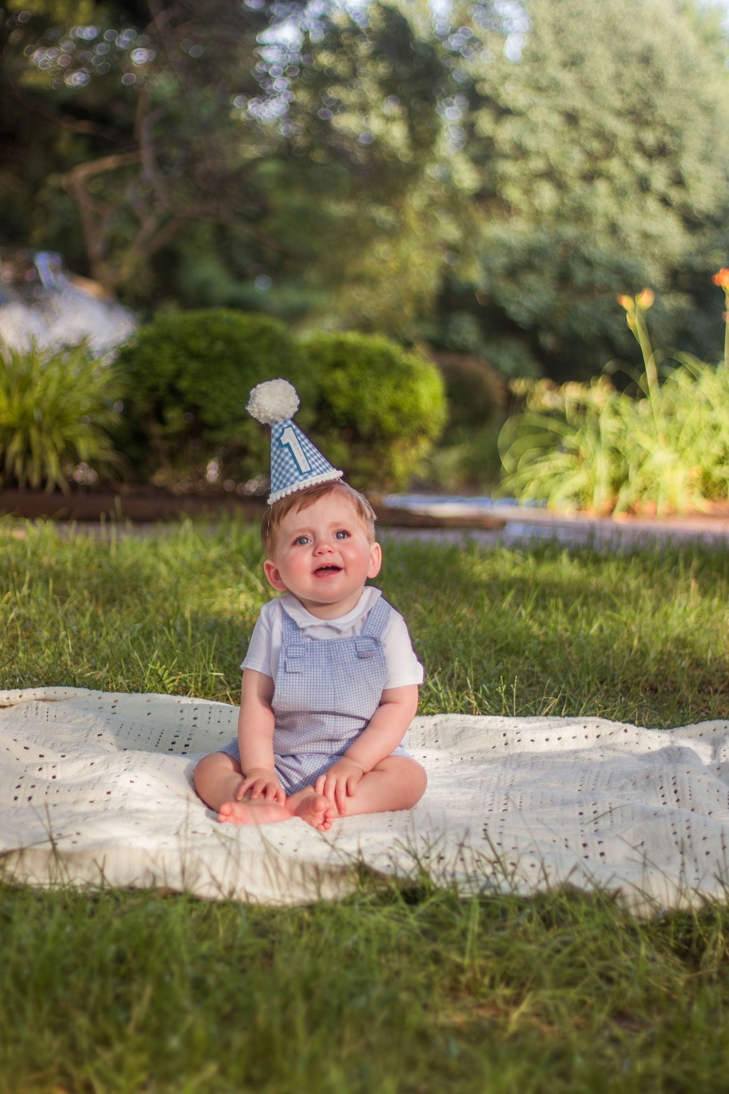 Happy baby wearing blue party hat sitting on white blanket in sunny green garden.
