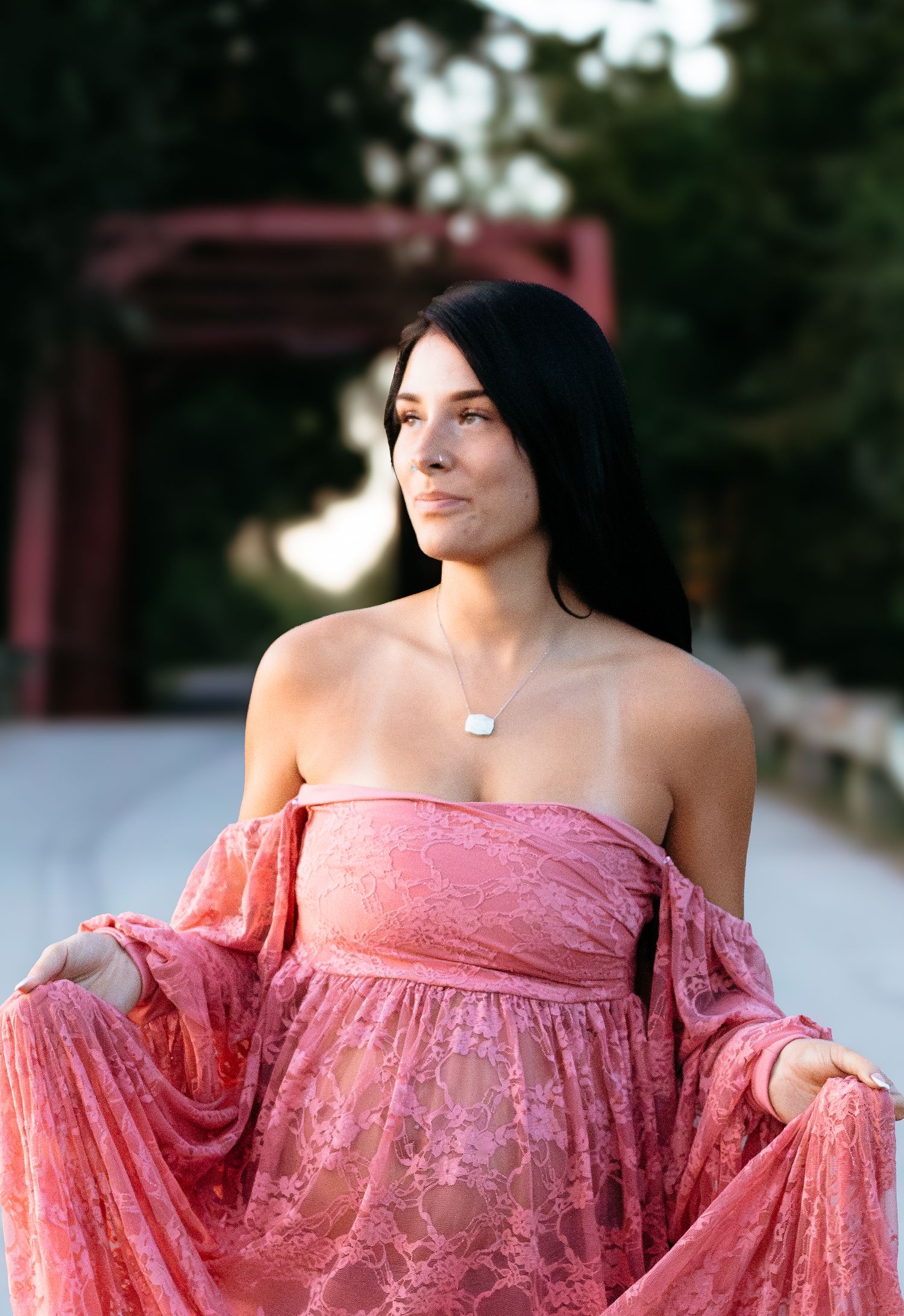 Pregnant woman in off-shoulder pink lace dress holding skirt open during outdoor maternity shoot.