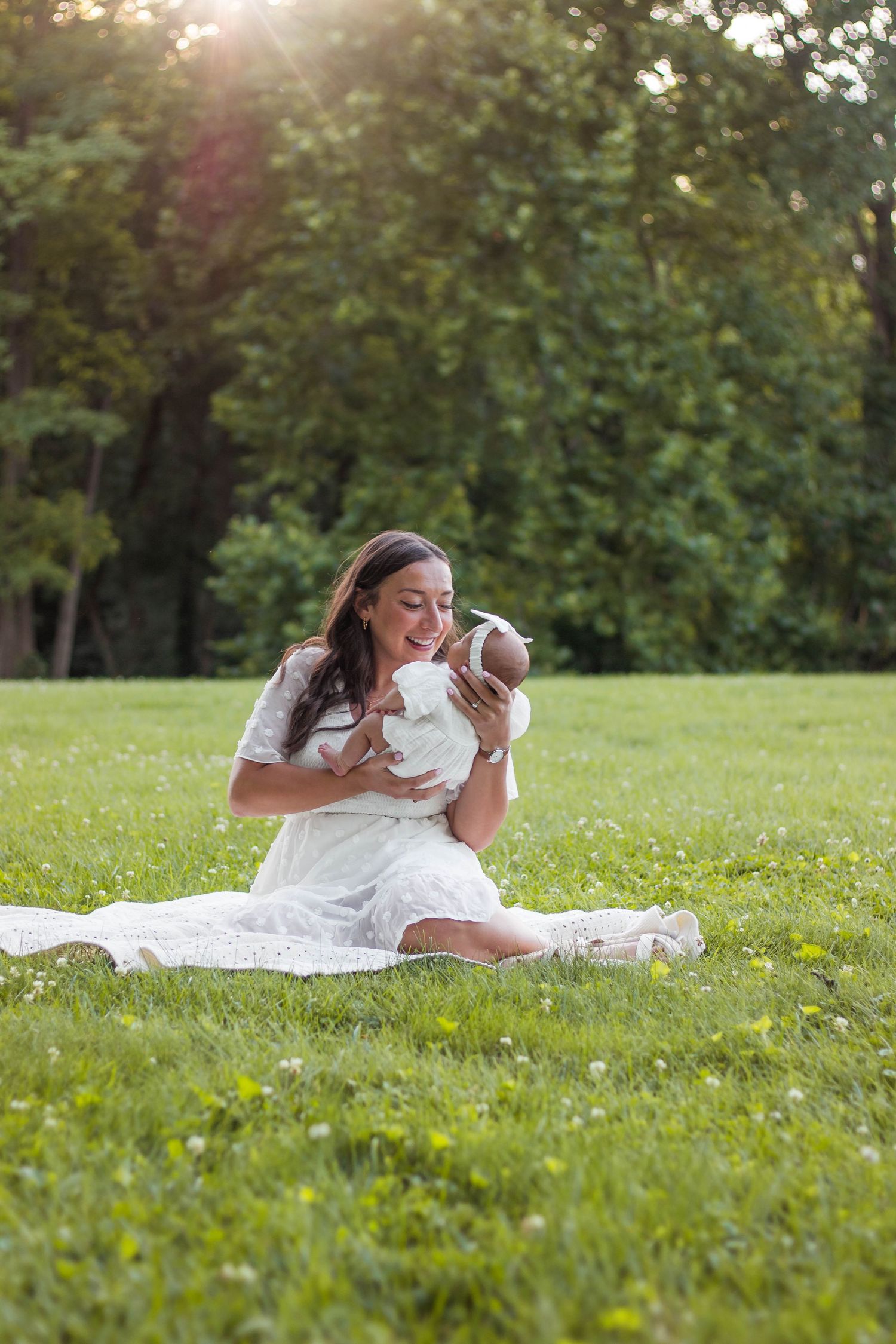 Mother in white dress sitting on grass holding newborn baby in sunny park.