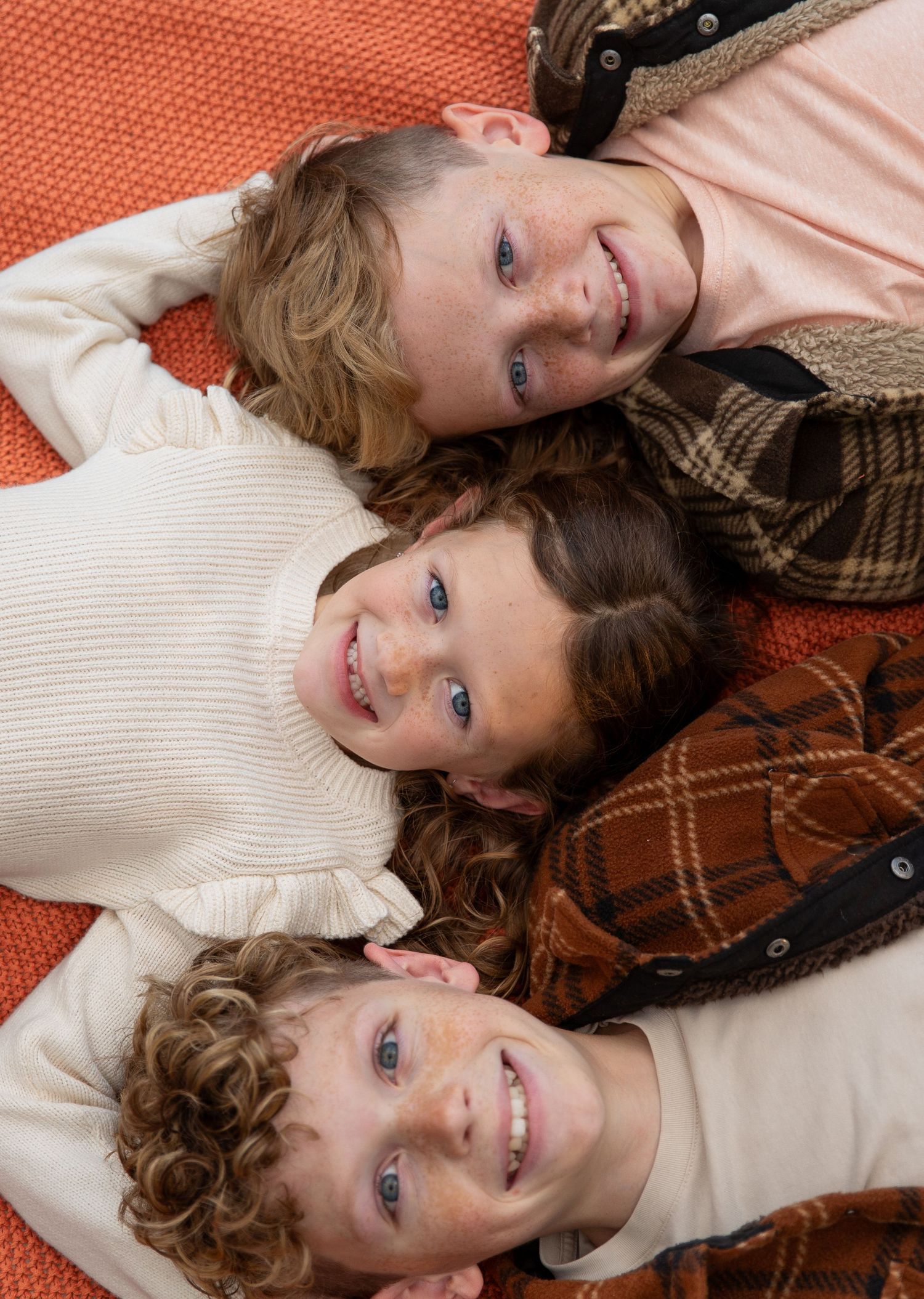 Three children lie on a brown plaid blanket wearing cozy sweaters while smiling at the camera- Rome GA Jessica M Cooper Photography LLC