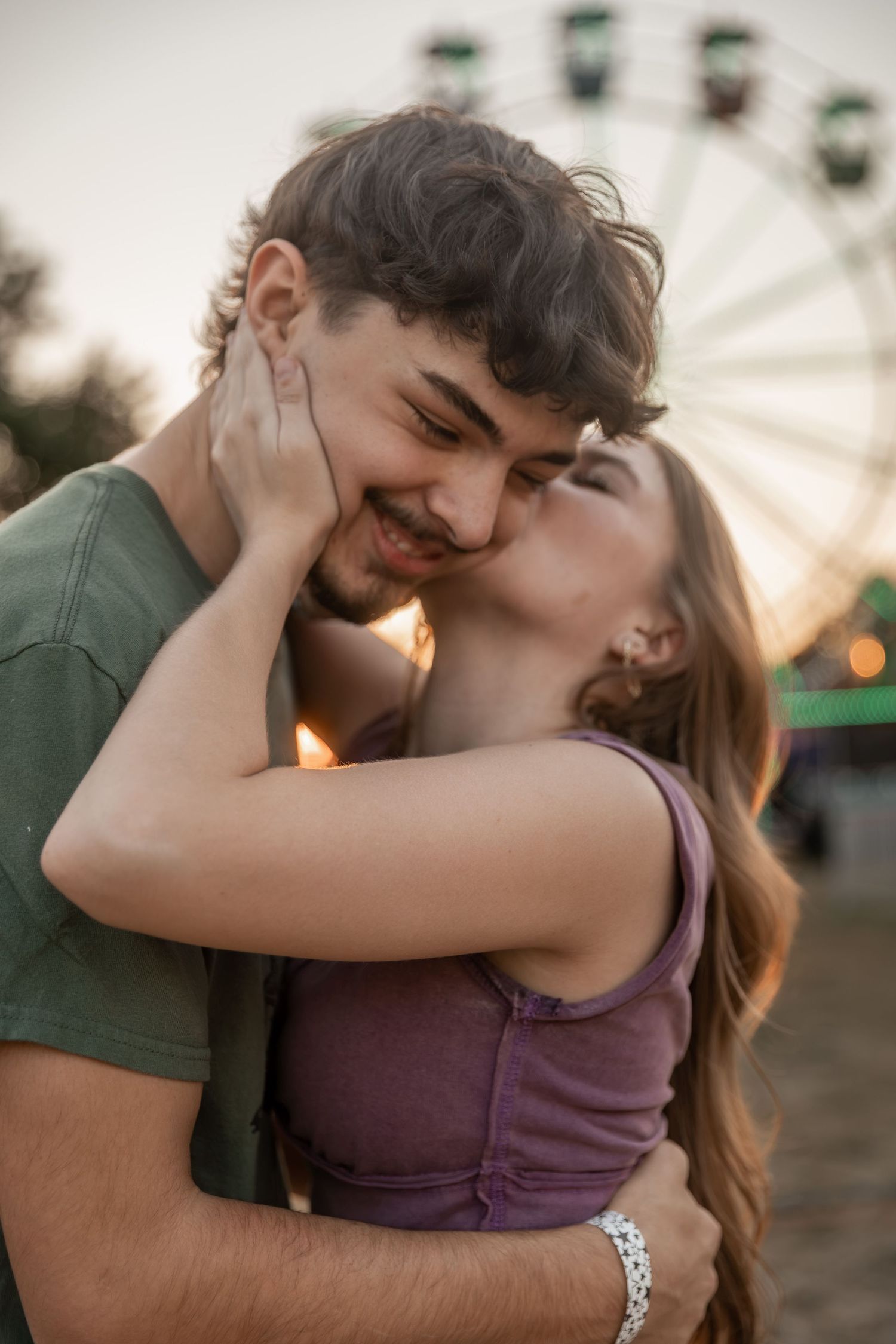 A couple sharing a tender kiss at a fairground with a Ferris wheel in the background at sunset.