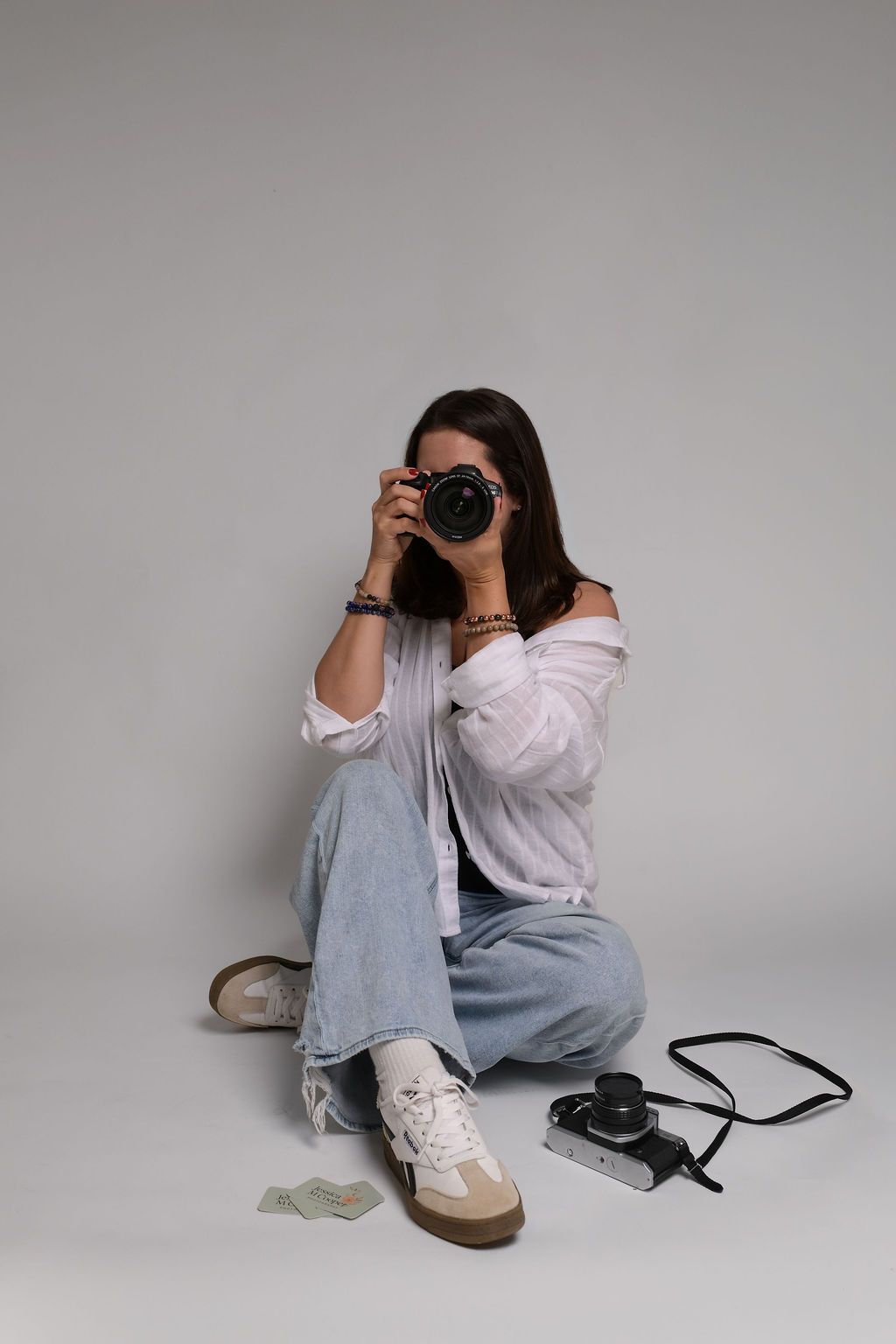 Young woman sitting on floor holding DSLR camera up to face, wearing white shirt and jeans, with vintage camera beside her.