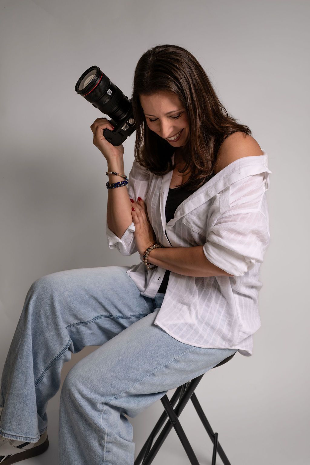 Young woman sitting on a stool holding a Canon DSLR camera, wearing white shirt and jeans, smiling in a studio setting.