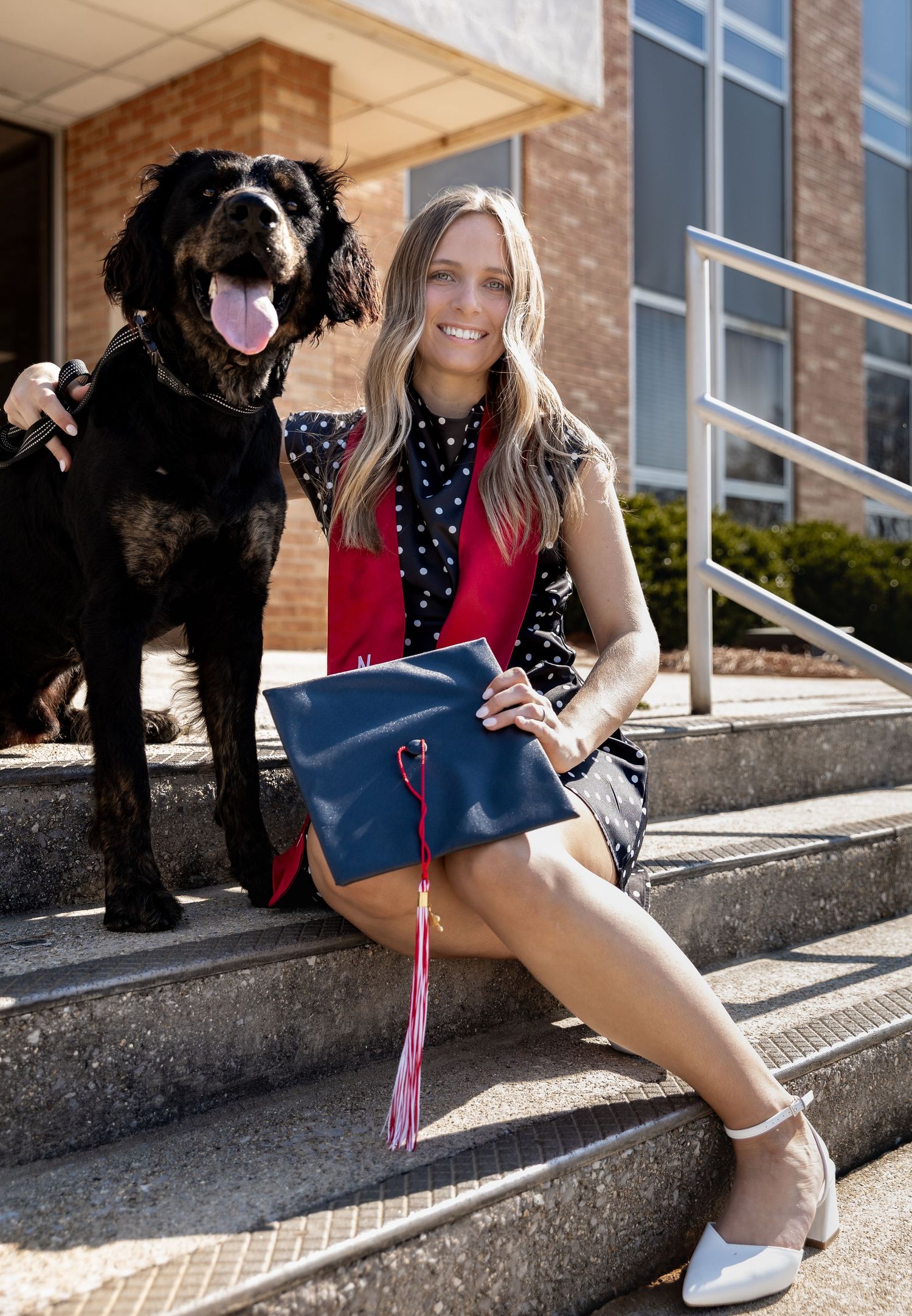 A smiling woman in graduation attire holds a navy cap on steps with a black dog beside her- College Graduation Jessica M Cooper Photography