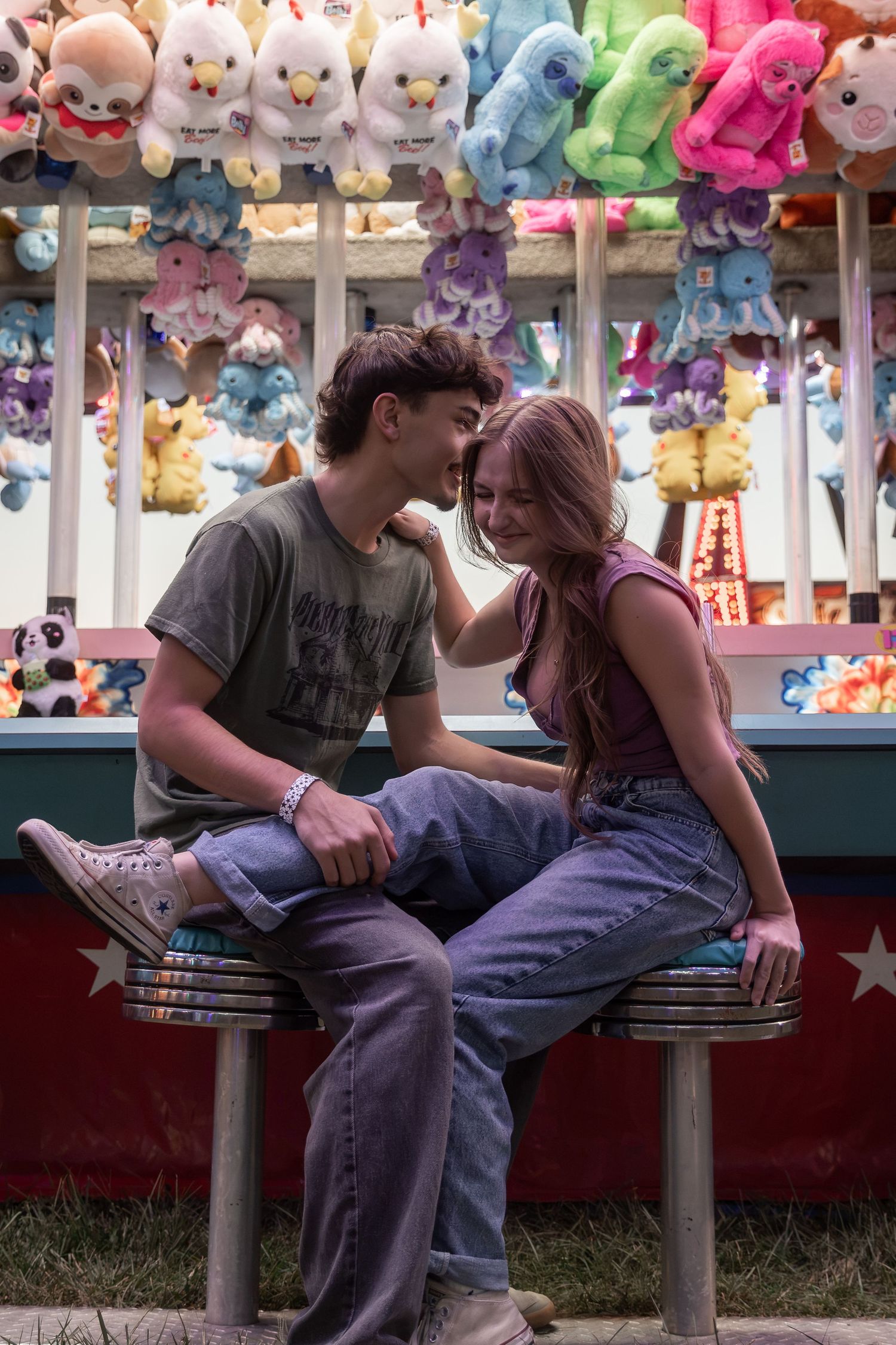 Young couple sharing a romantic moment at a colorful carnival game booth surrounded by stuffed animal prizes.