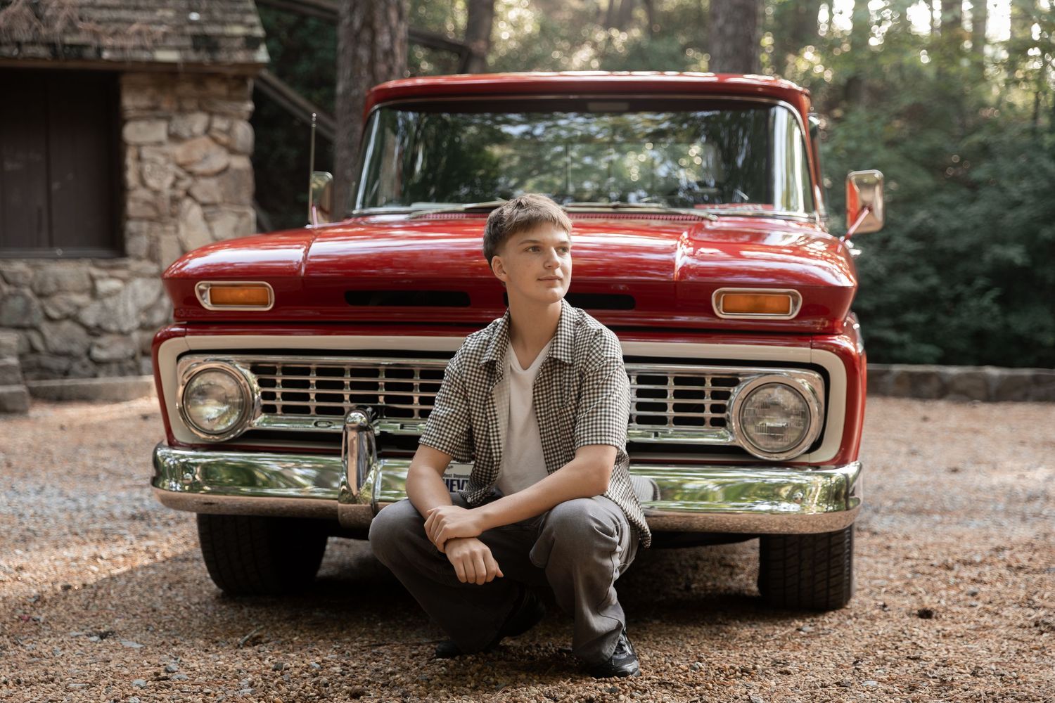 Young boy sitting in front of a classic red vintage pickup truck on a gravel driveway surrounded by trees.