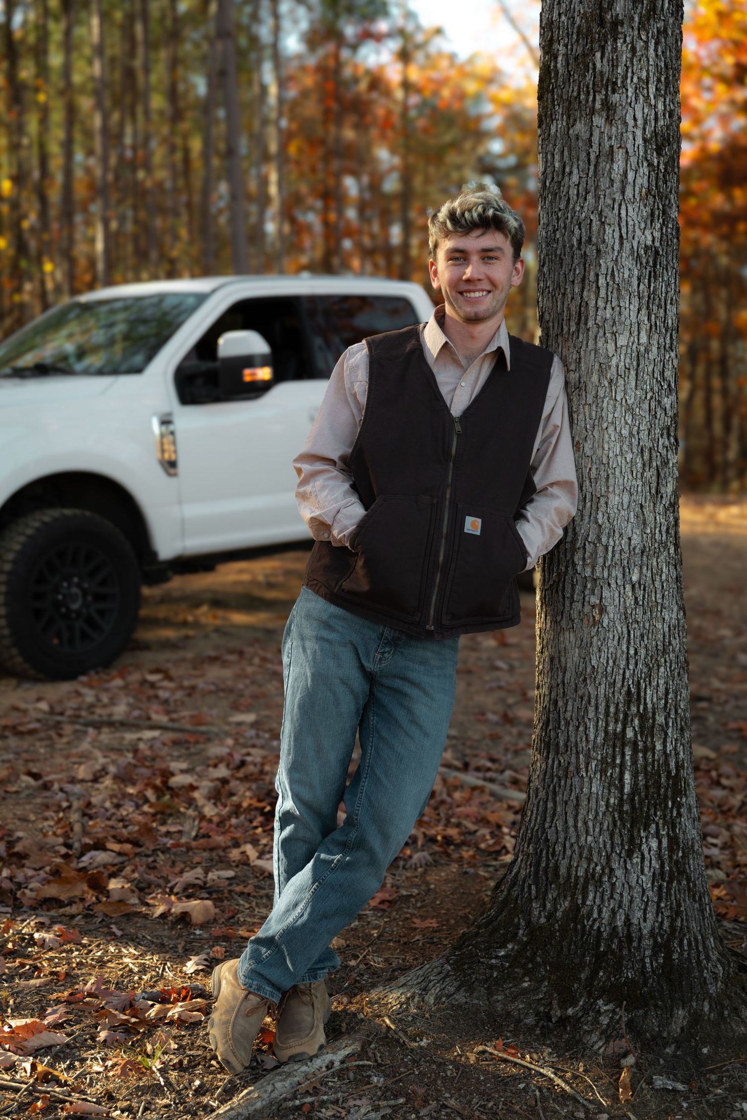 Young man in Carhartt vest and jeans leaning against tree in autumn forest with white pickup truck behind him- White, GA Senior portraits