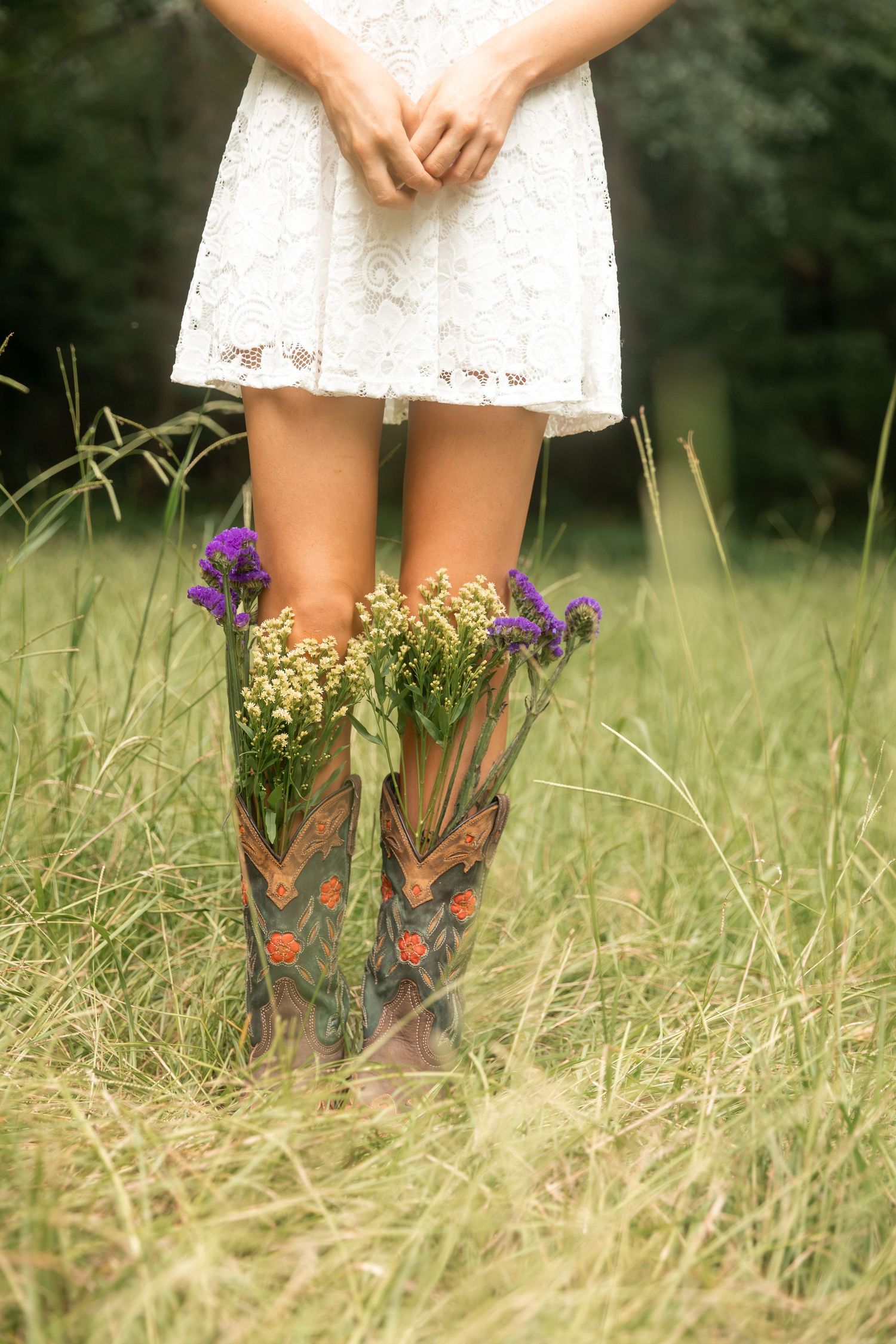 Woman in white lace dress wearing decorative cowboy boots filled with wildflowers standing in tall grass field.