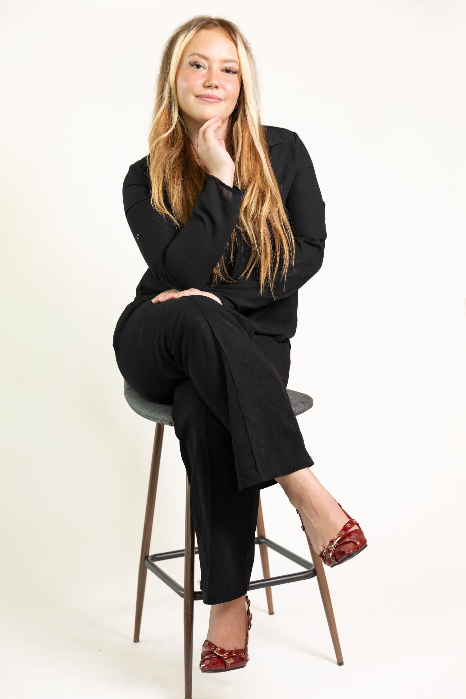 Young woman in black outfit and red flats sitting on a stool with hand on chin against white background. Branding session Acworth, GA
