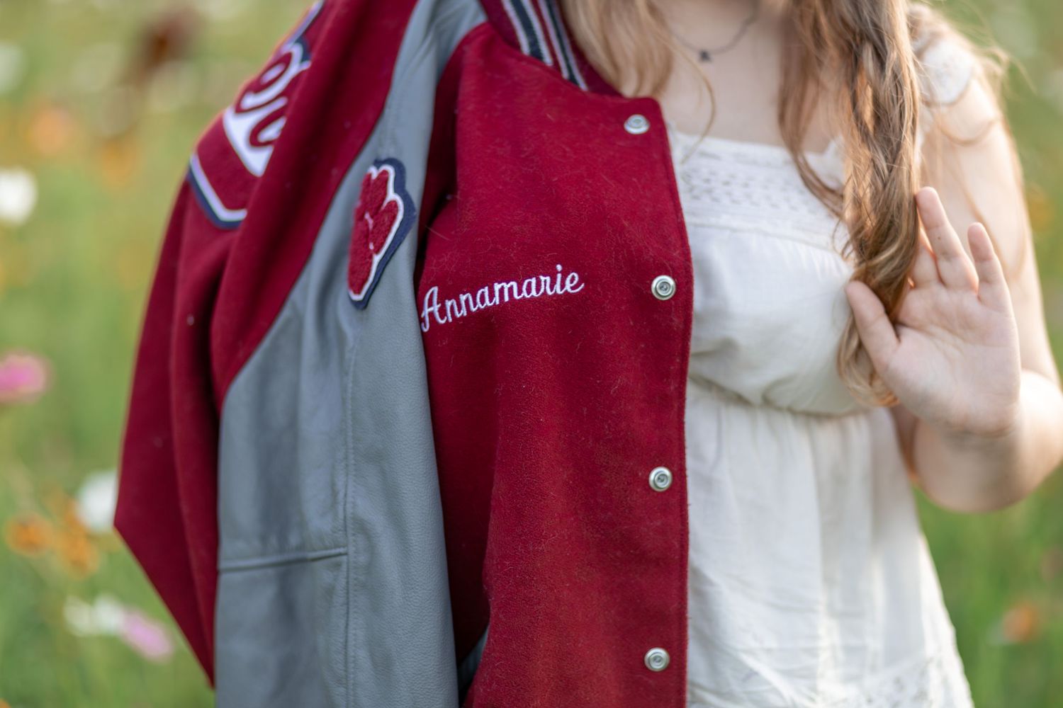 A red and gray varsity letterman jacket with the name Annamarie embroidered on it, held by a young woman outdoors.