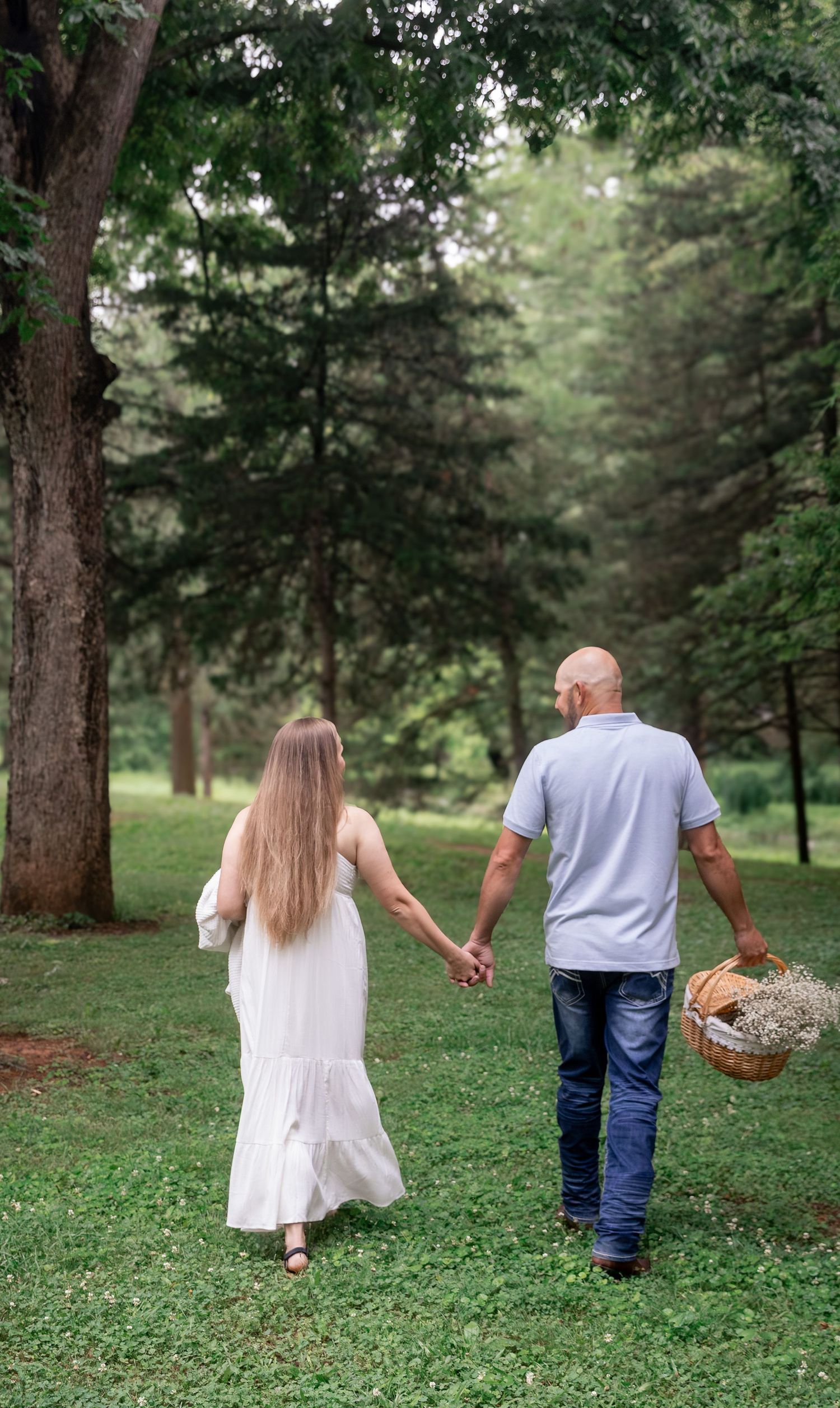 A couple walks hand-in-hand through a lush green park, the man carrying a wicker basket while the woman wears a flowing white dress.