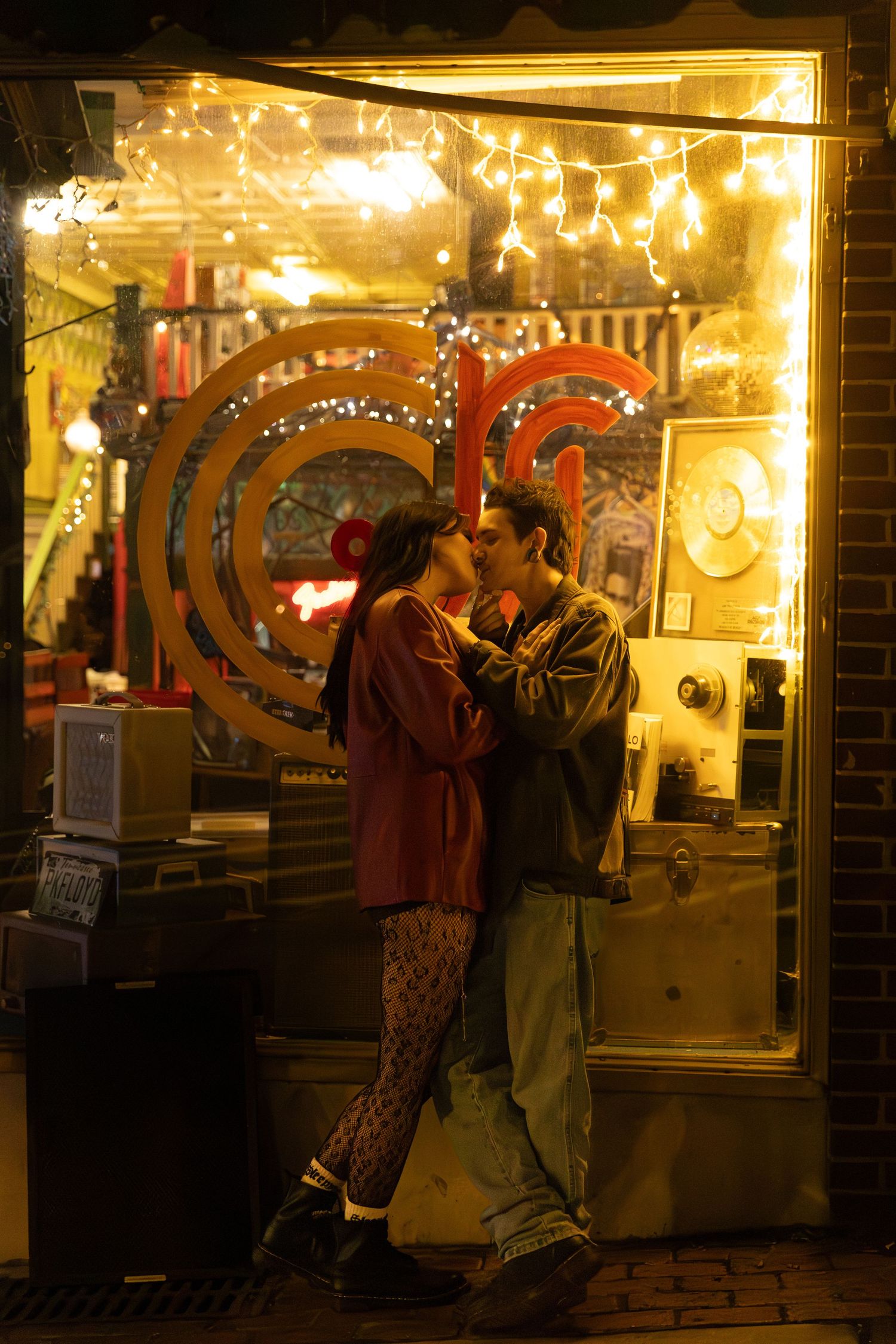 Couple sharing a romantic kiss outside a warmly lit shop with fairy lights and gold records at night. Downtown Rome, GA