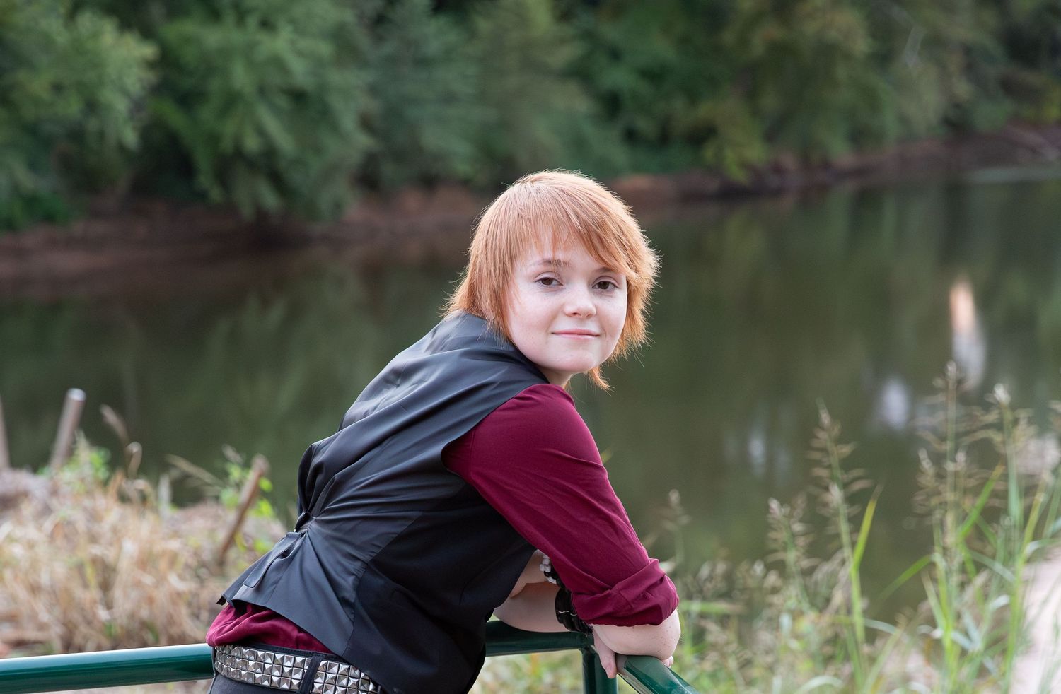 Red-haired child smiling over shoulder, leaning on green railing near a calm river with lush green trees downtown Rome, GA