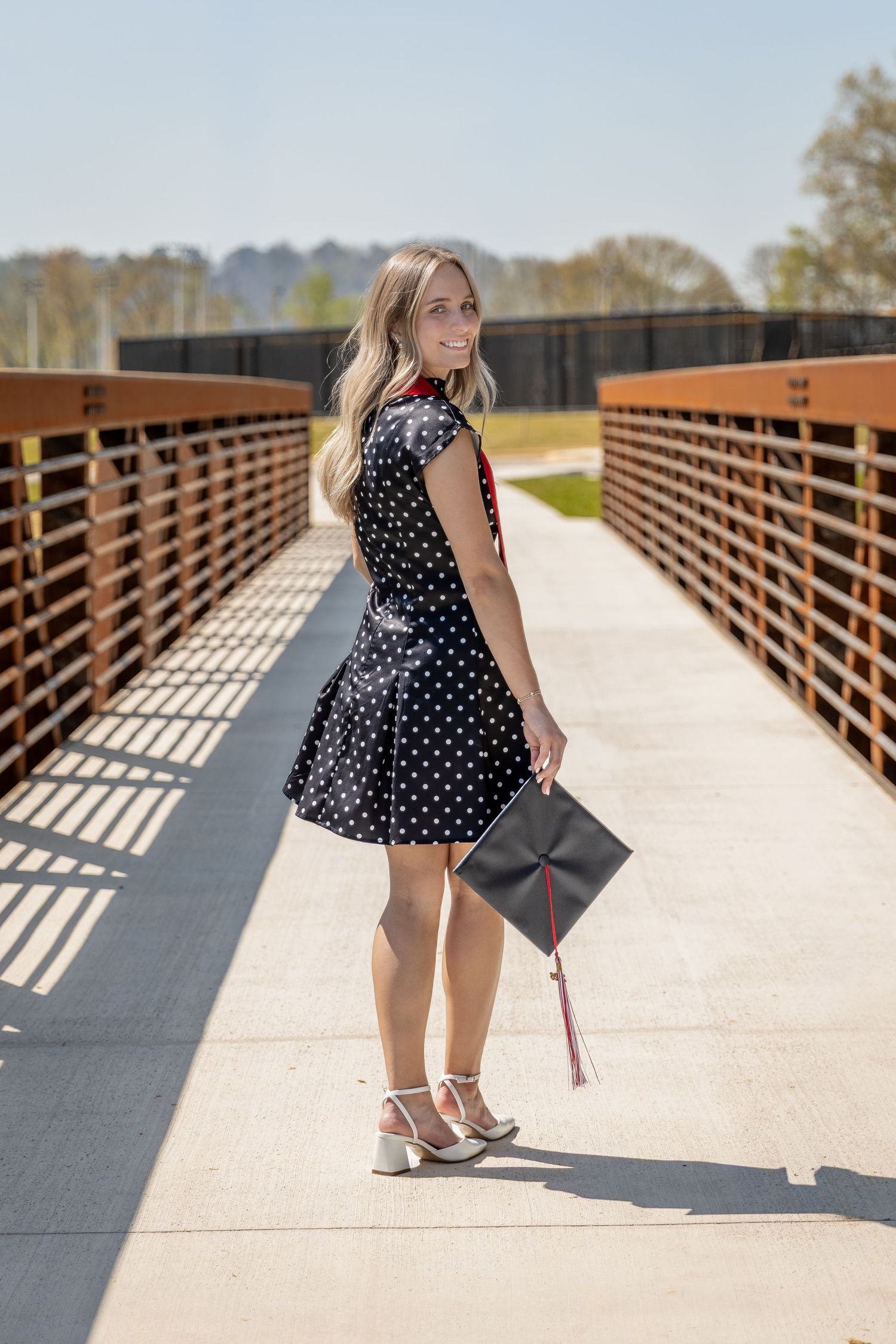 Smiling graduate in polka dot dress holding cap on sunny bridge, celebrating graduation day outdoors Northwest GA