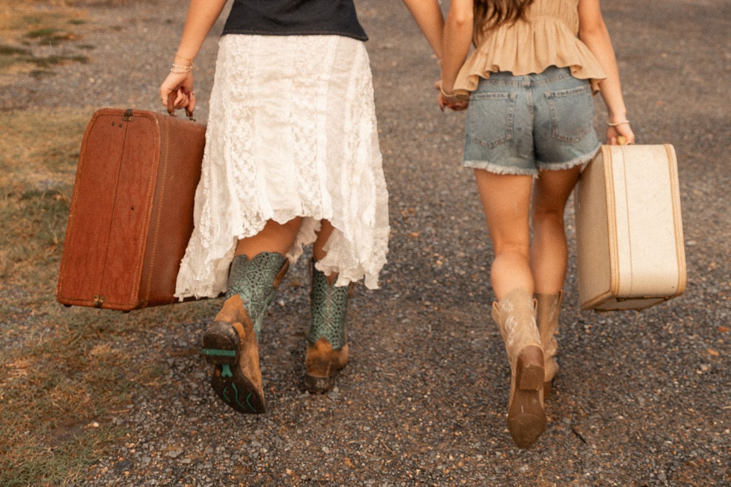 Two women in boho outfits and cowboy boots walk away carrying vintage suitcases on a gravel road.