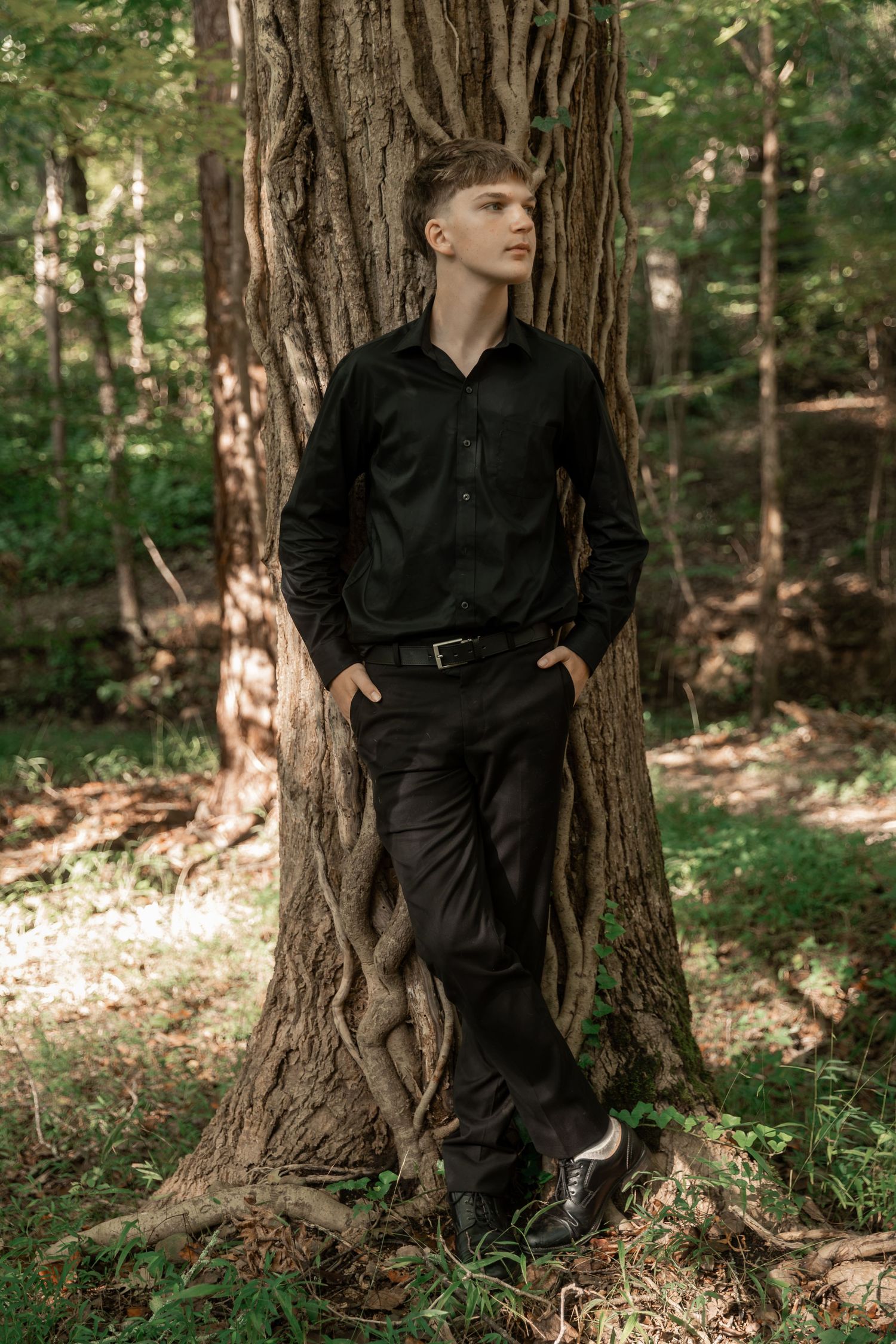 Young man in black outfit leaning against large tree trunk in forest setting.