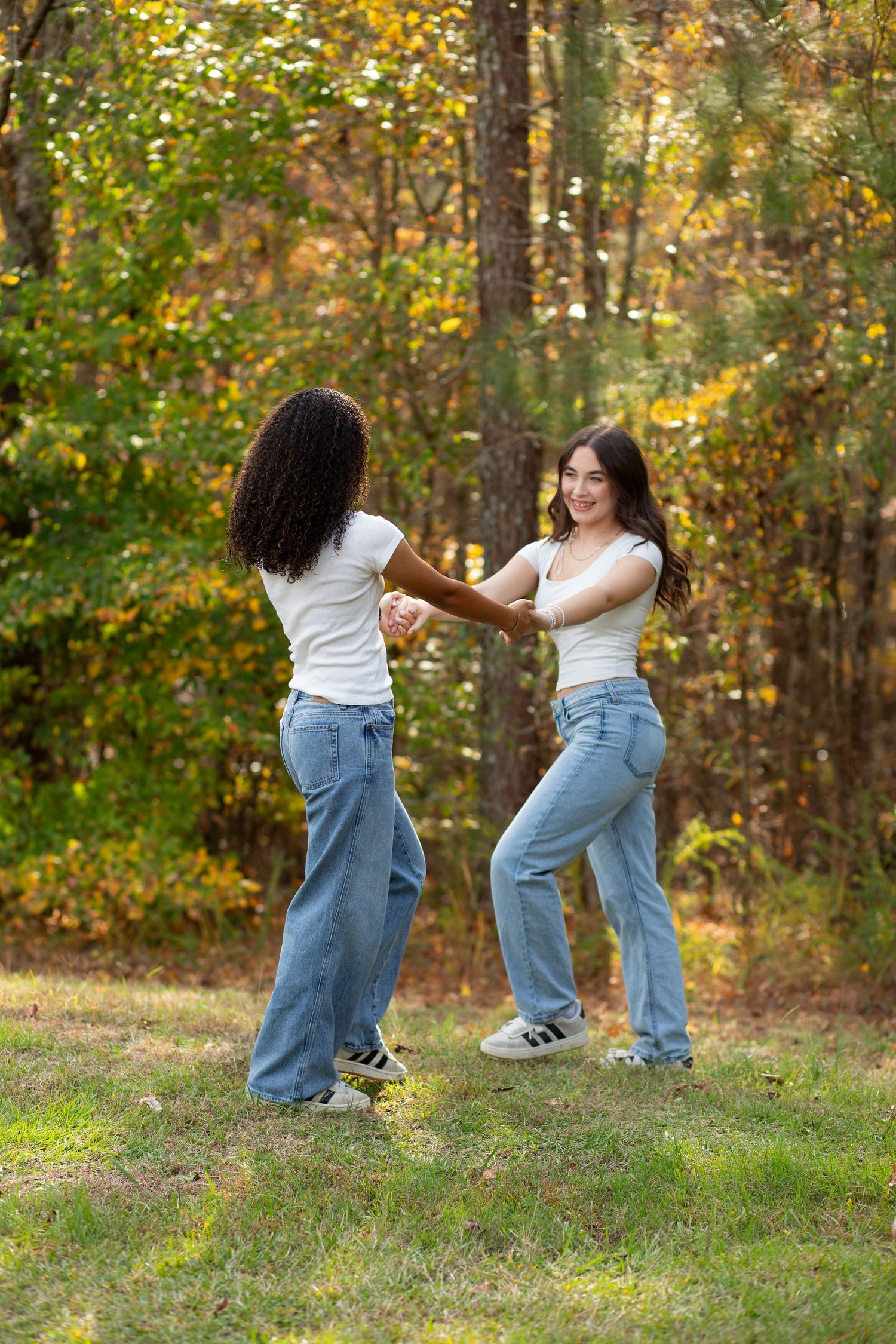 Two young women in white tops and jeans hold hands while dancing outdoors among colorful autumn trees.