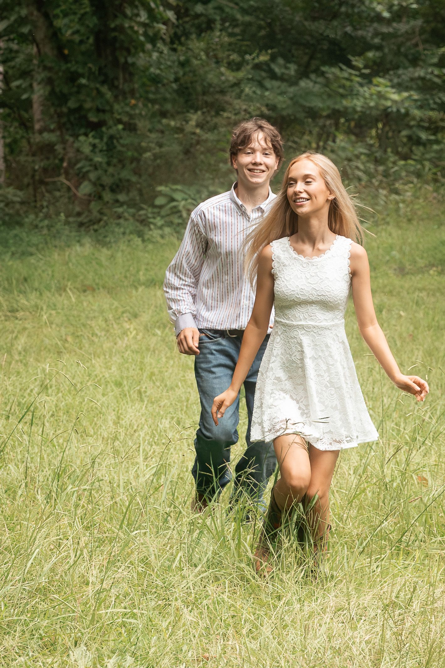 A smiling young couple walks through a lush green meadow, the woman in a white lace dress and brown boots. Rome, GA
