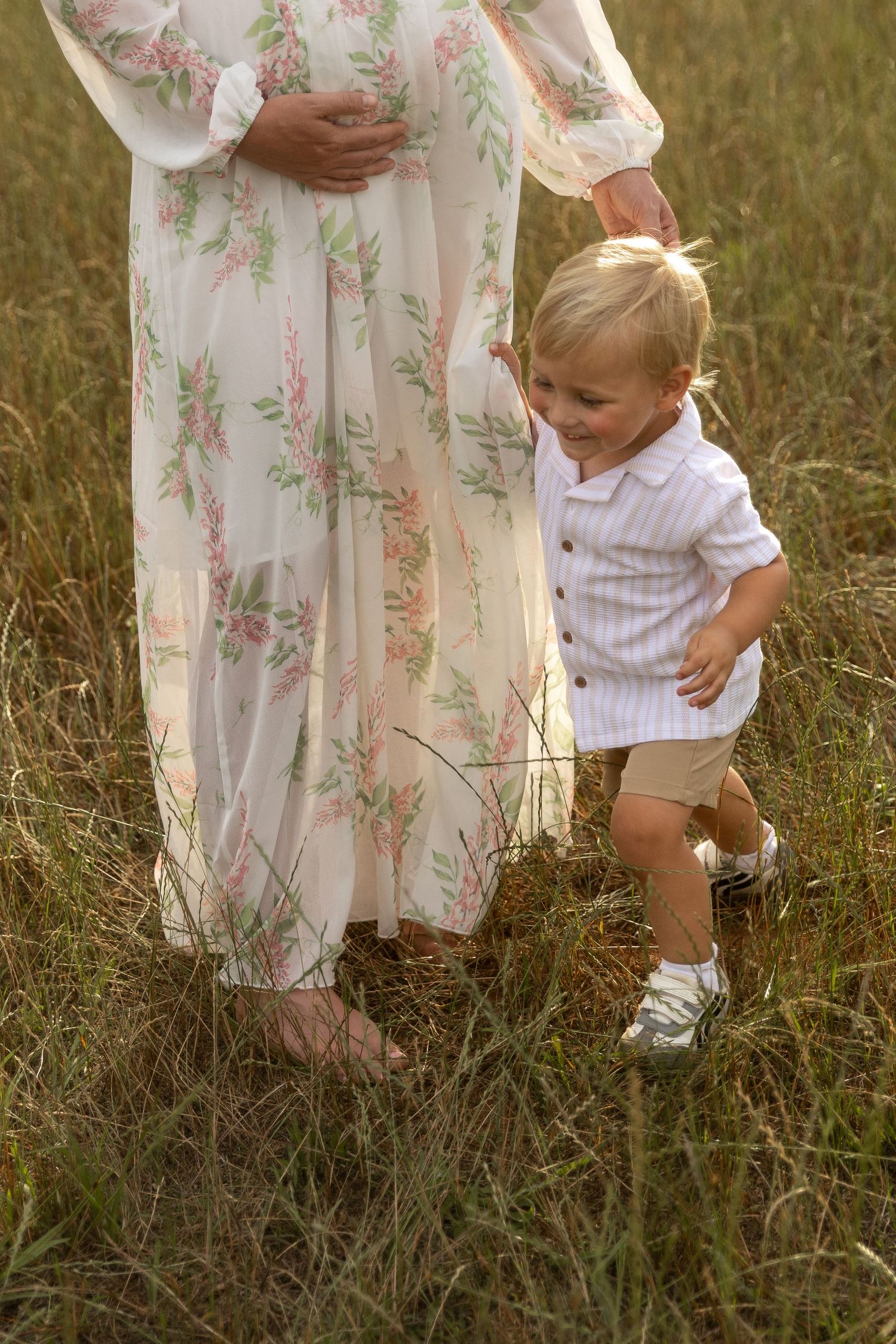 A smiling toddler boy in a white shirt stands beside his pregnant mothers floral dress in a grassy field.